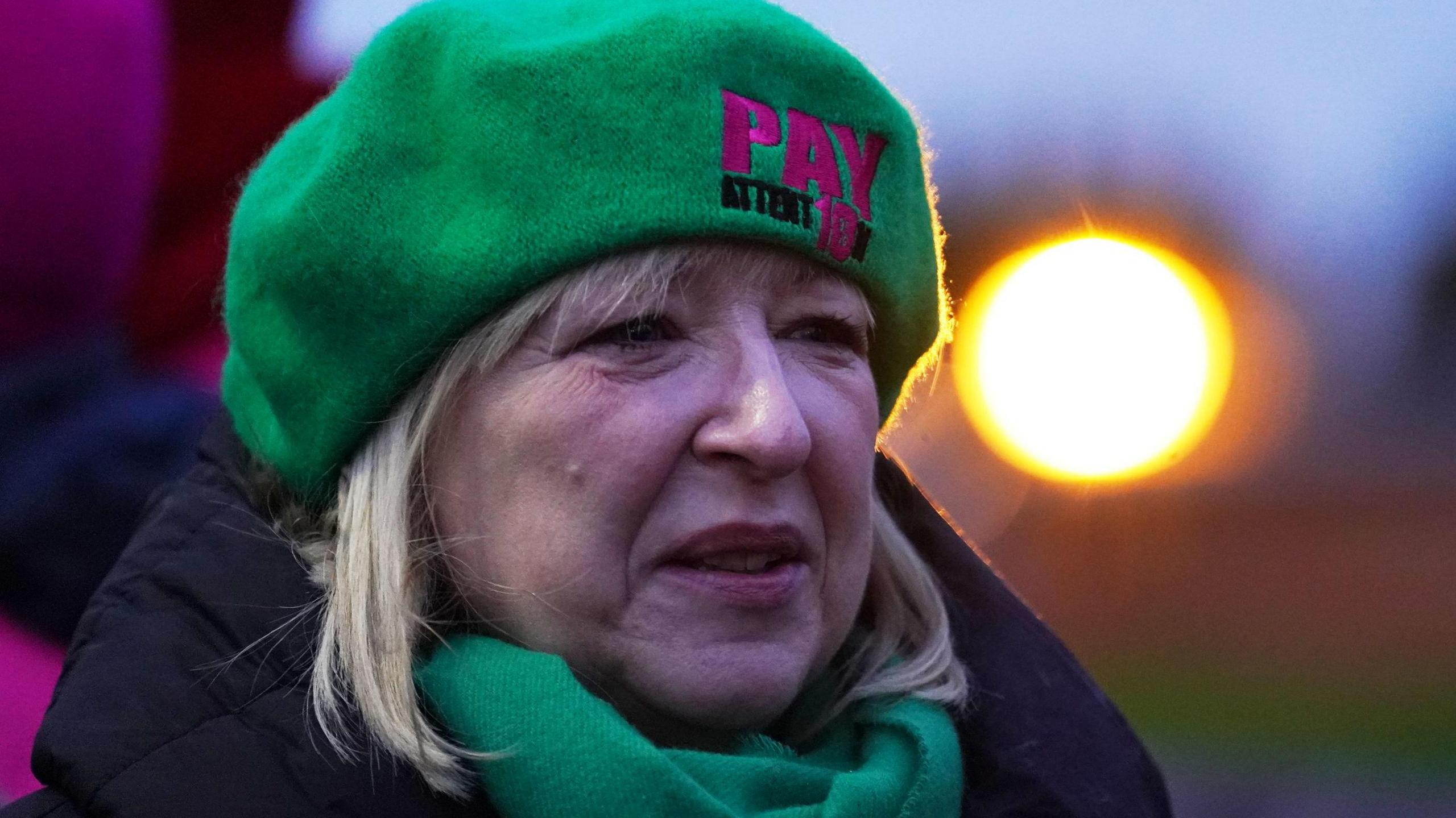 A woman with shoulder length blonde hair looks to the side slightly. She wears a green hat and scarf and a black jacket