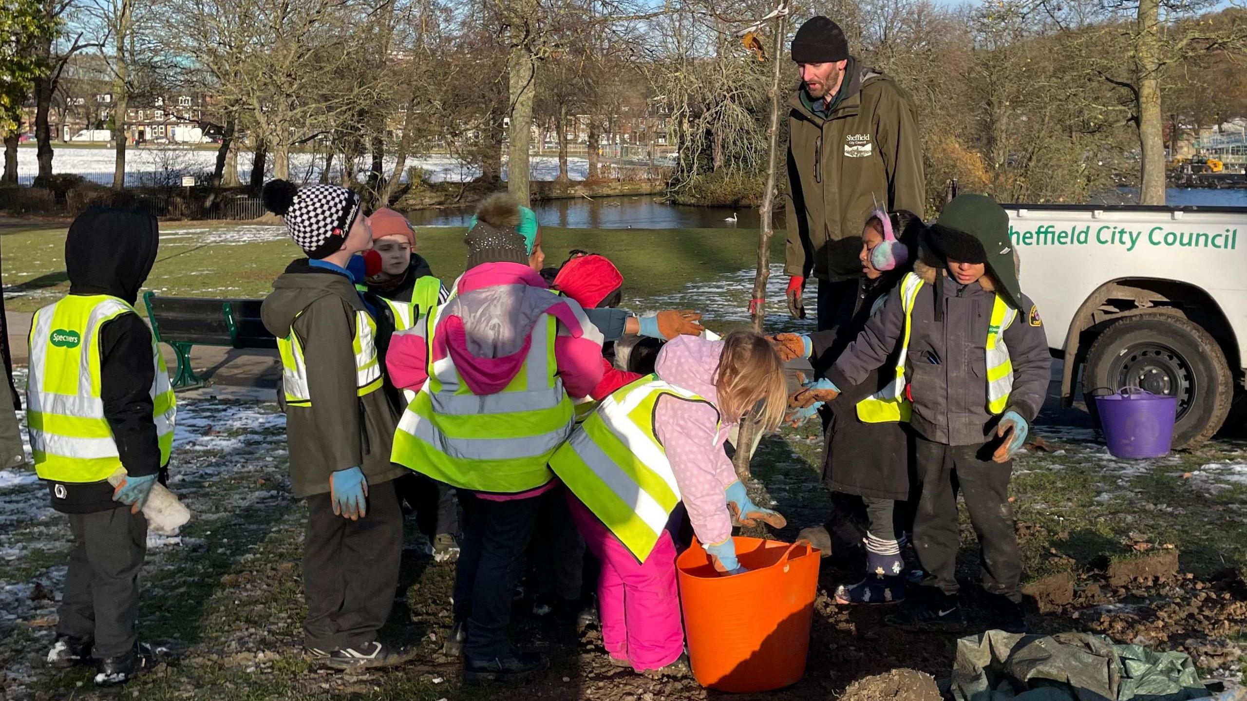 The children are gathered around the sapling, listening to a man from the council who's talking. One girl is reaching into an orange bucket filled with food for the tree.