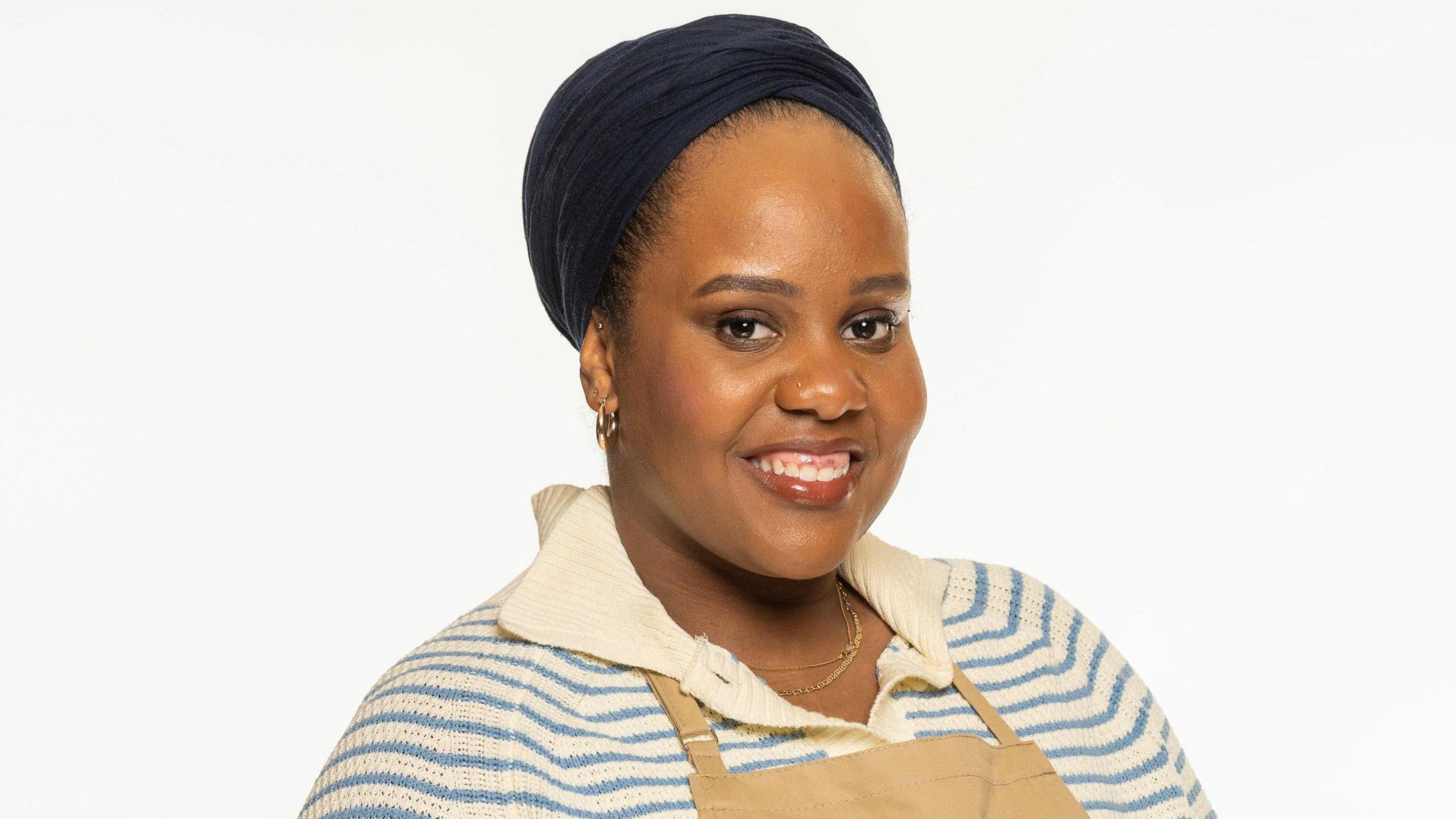 Illiyin Morrison standing in front of a white background and is wearing a blue and white striped top with a brown apron. 