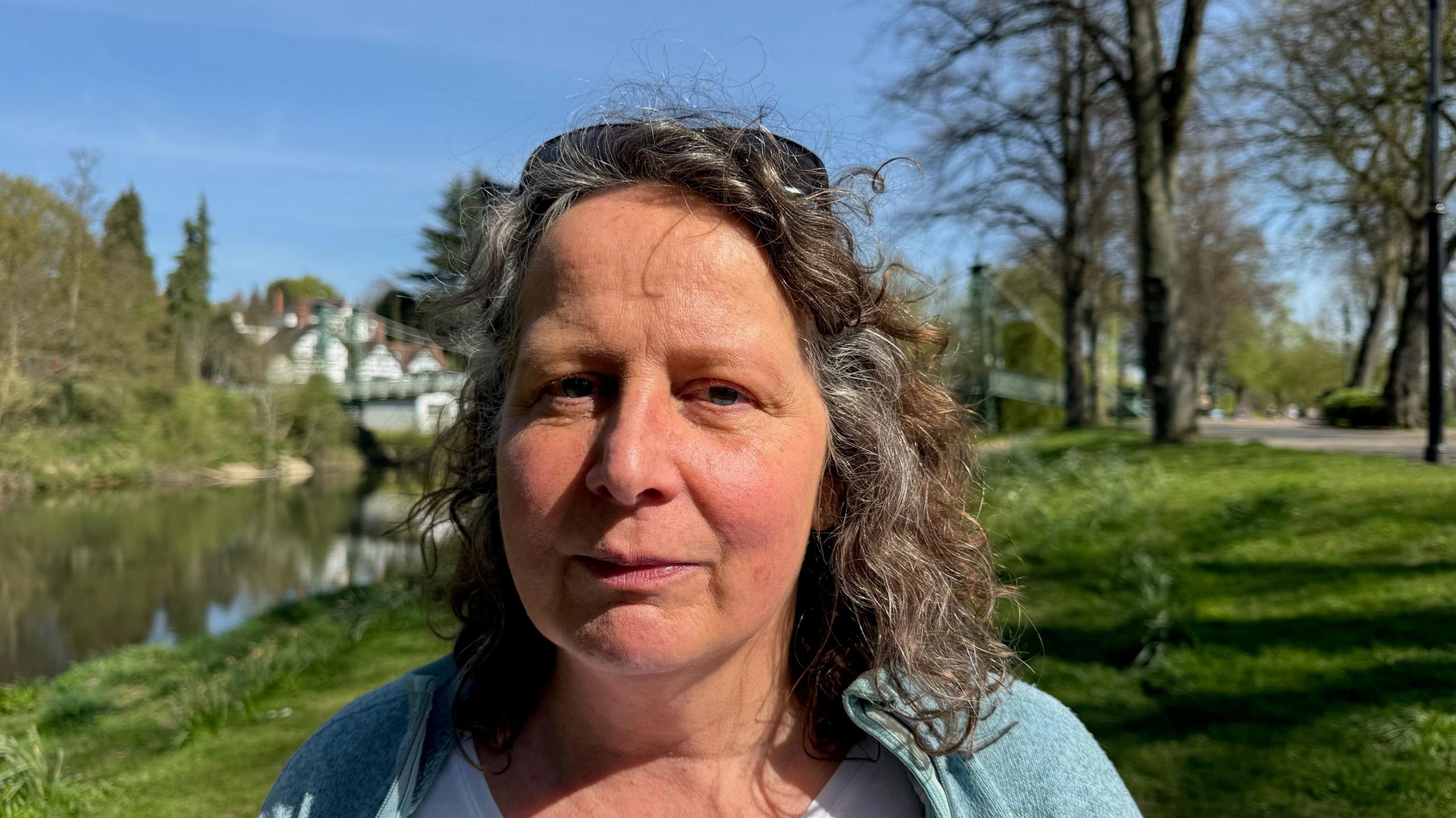 Dr Alison Caffyn is a white woman in her 50s. She is standing in front of the River Severn in Shrewsbury. She has shoulder length hair and she is wearing a blue cardigan.