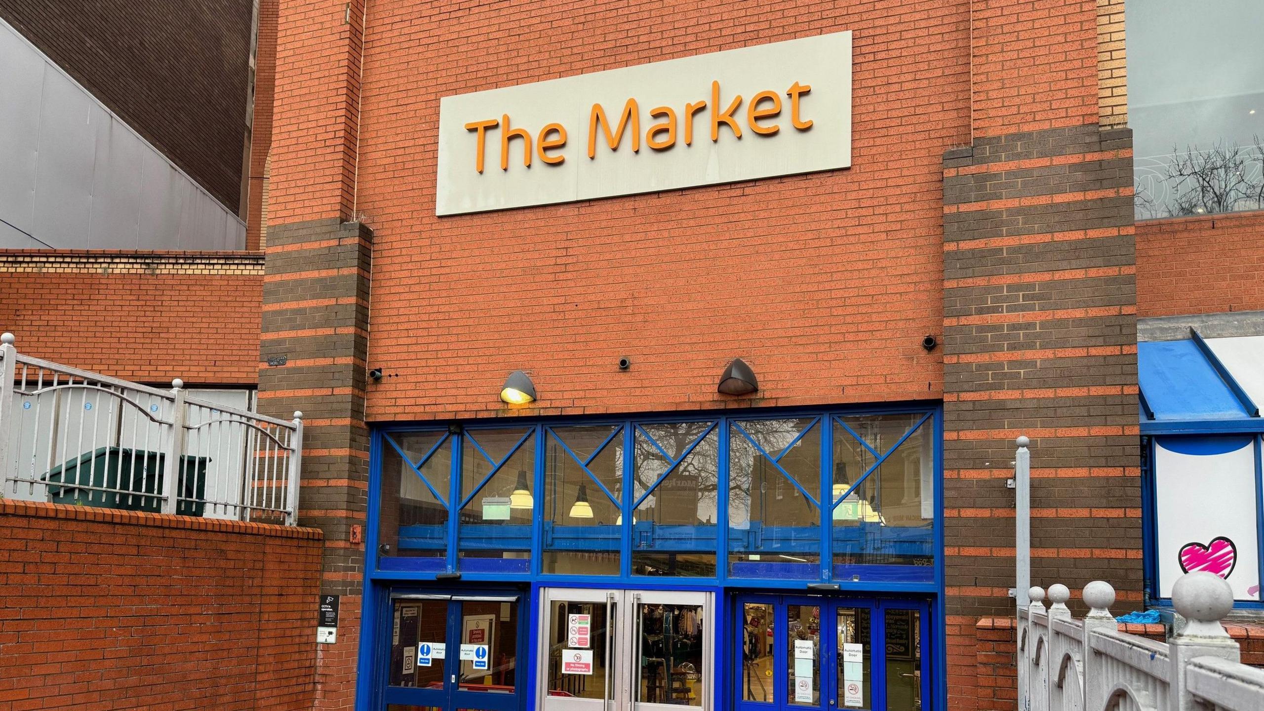 The entrance to Hanley Indoor Market, with a large sign above blue folding doors which reads "The Market" and sets of railing on either side a ramp down to the market.