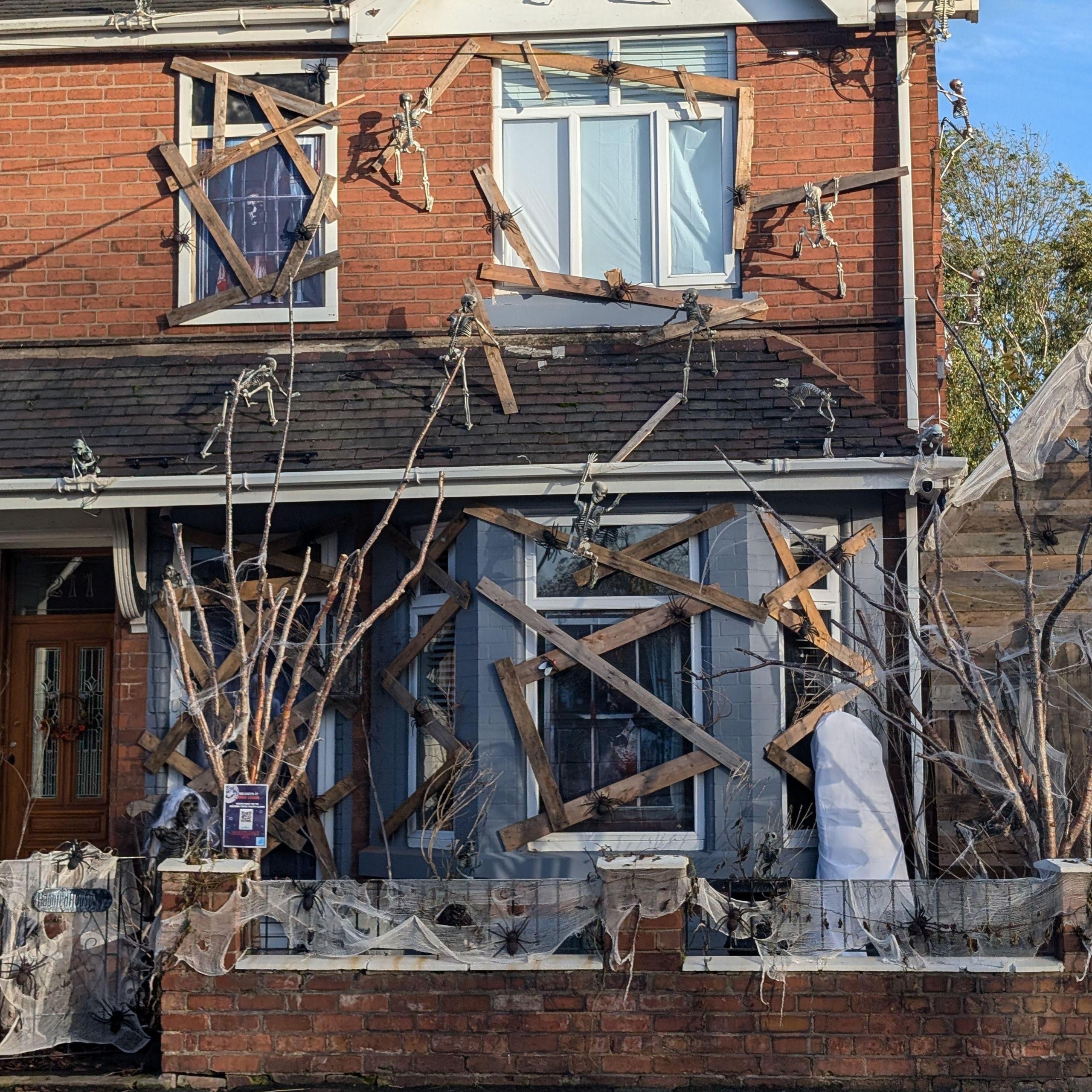 The front of a semi-detached house with Halloween decorations. Wooden boards have been placed over windows and doors, while model skeletons can be seen climbing the building.