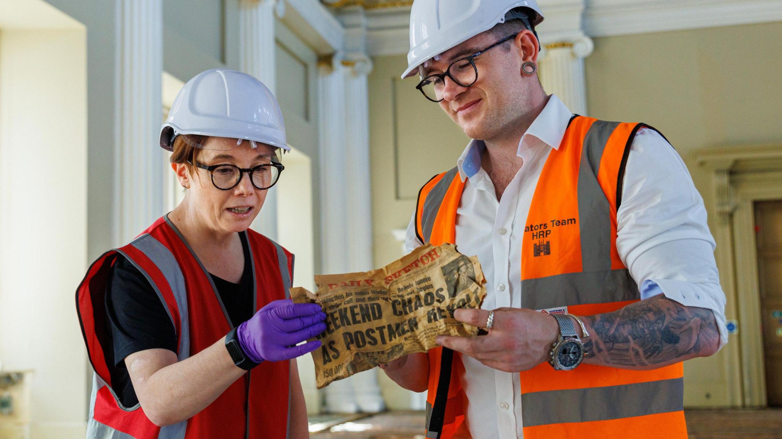 One woman and one man wearing orange hi-vis and white hard hats holding the Daily Sketch which said Weekend Chaos as Postmen Revolt. The man has tattoos on his left arm and is wearing a silver watch and earring. The lady is wearing a plastic purple glove and has black glasses on.