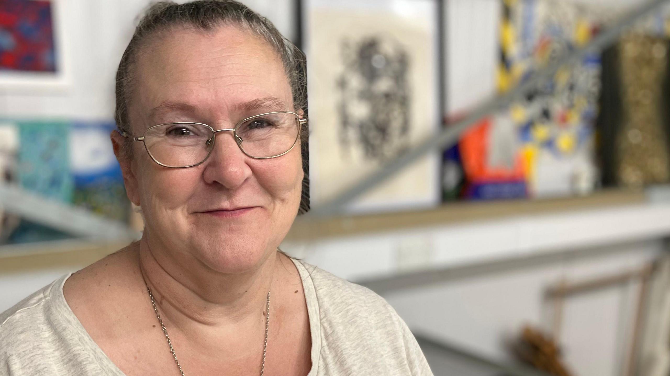 A woman smiles at the camera in a head and shoulders image. She is wearing a beige scoop-necked tshirt, thin gold chain necklace and silver framed spectacles. Her dark blonde hair is tied back and scraped away from her face. The blurred background is made up of framed artwork propped on a shelf
