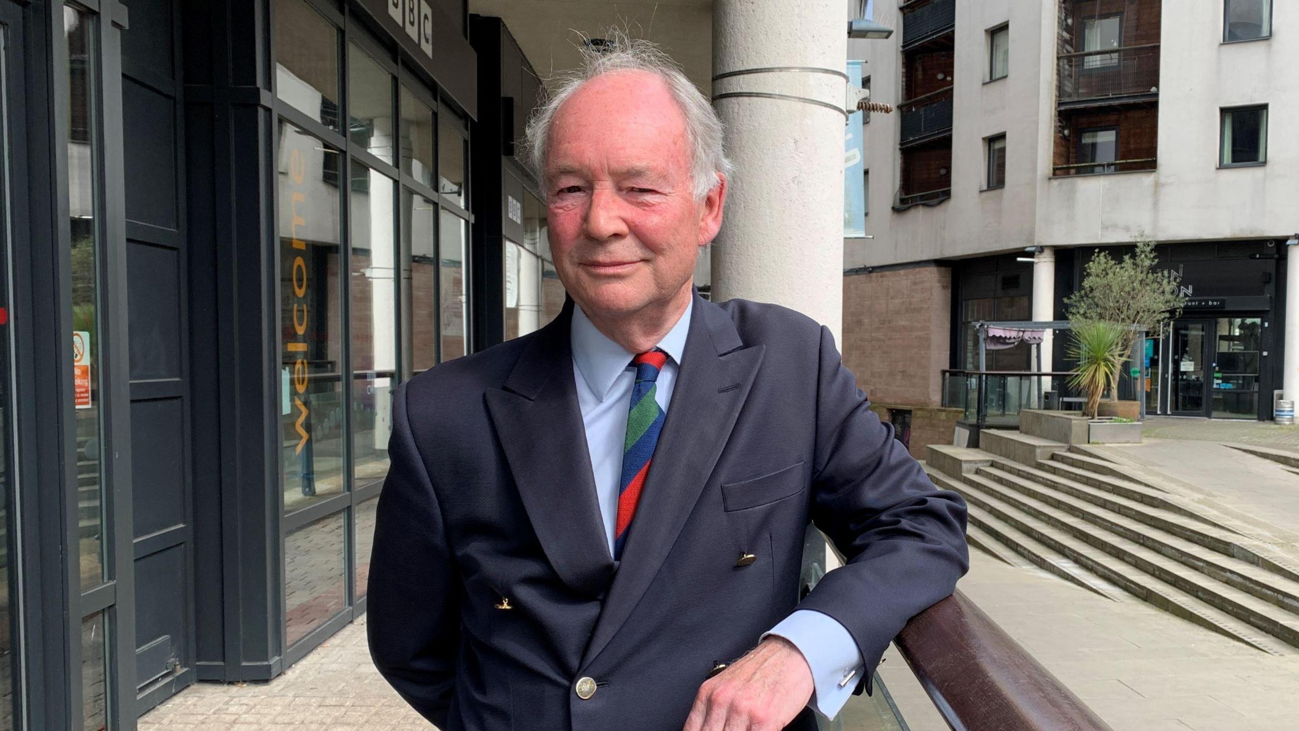 Philip Seccombe stood outside a white building with glass frontage wearing a dark blue suit and a multi-coloured tie