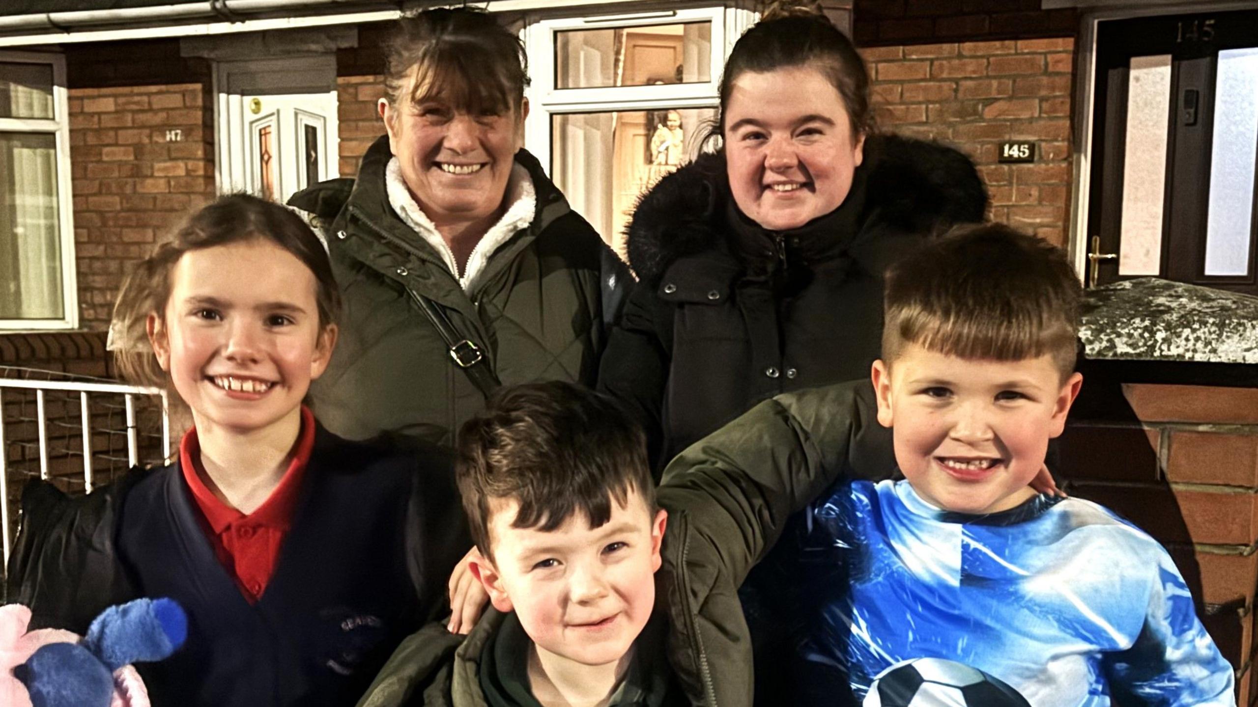 three children and two women smiling for the camera. One of the children is wearing a football themed tracksuit, one is wearing a thick rain coat, and the other is wearing a school uniform.