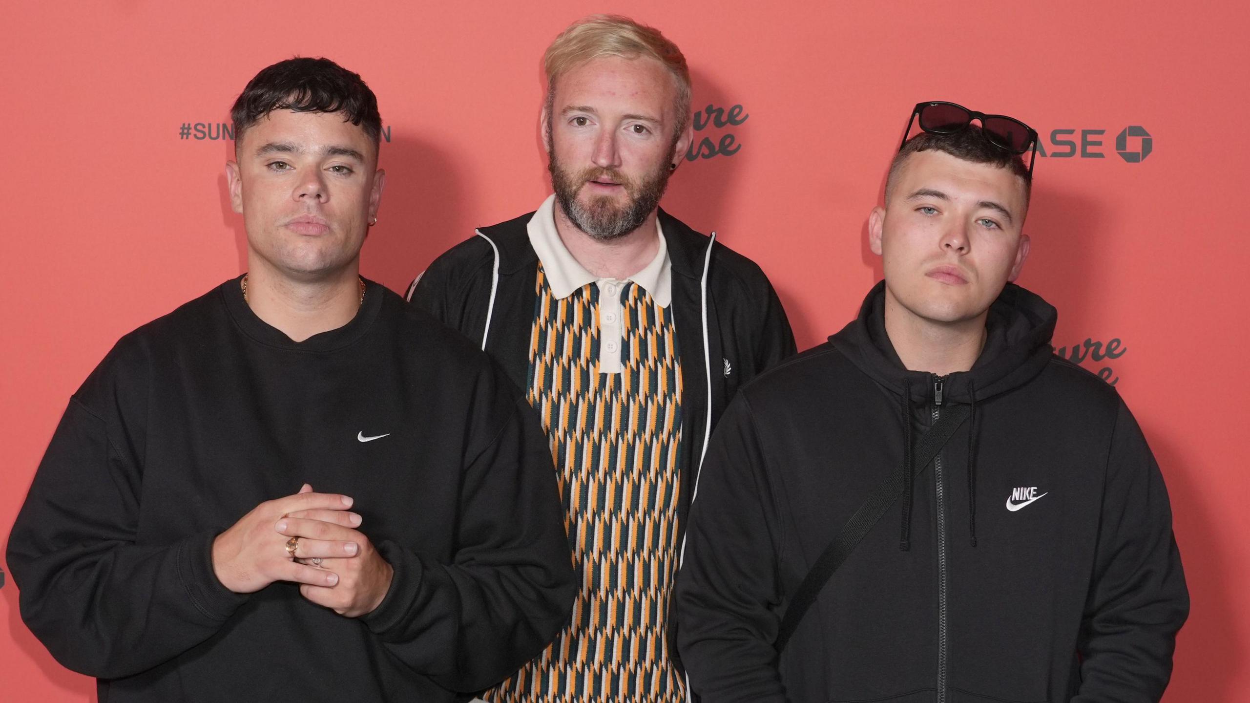 Mo Chara, left, is wearing a black sweatshirt and looking directly at the camera. He had short brown hair. DJ Provai, middle, is wearing a yellow and green patterned polo top and a black zipper jacket. He is looking directly at the camera and has a beard and ginger hair. Moglai Bap, is wearing a black zip up coat. He is wearing sunglasses on his head and has short brown hair.