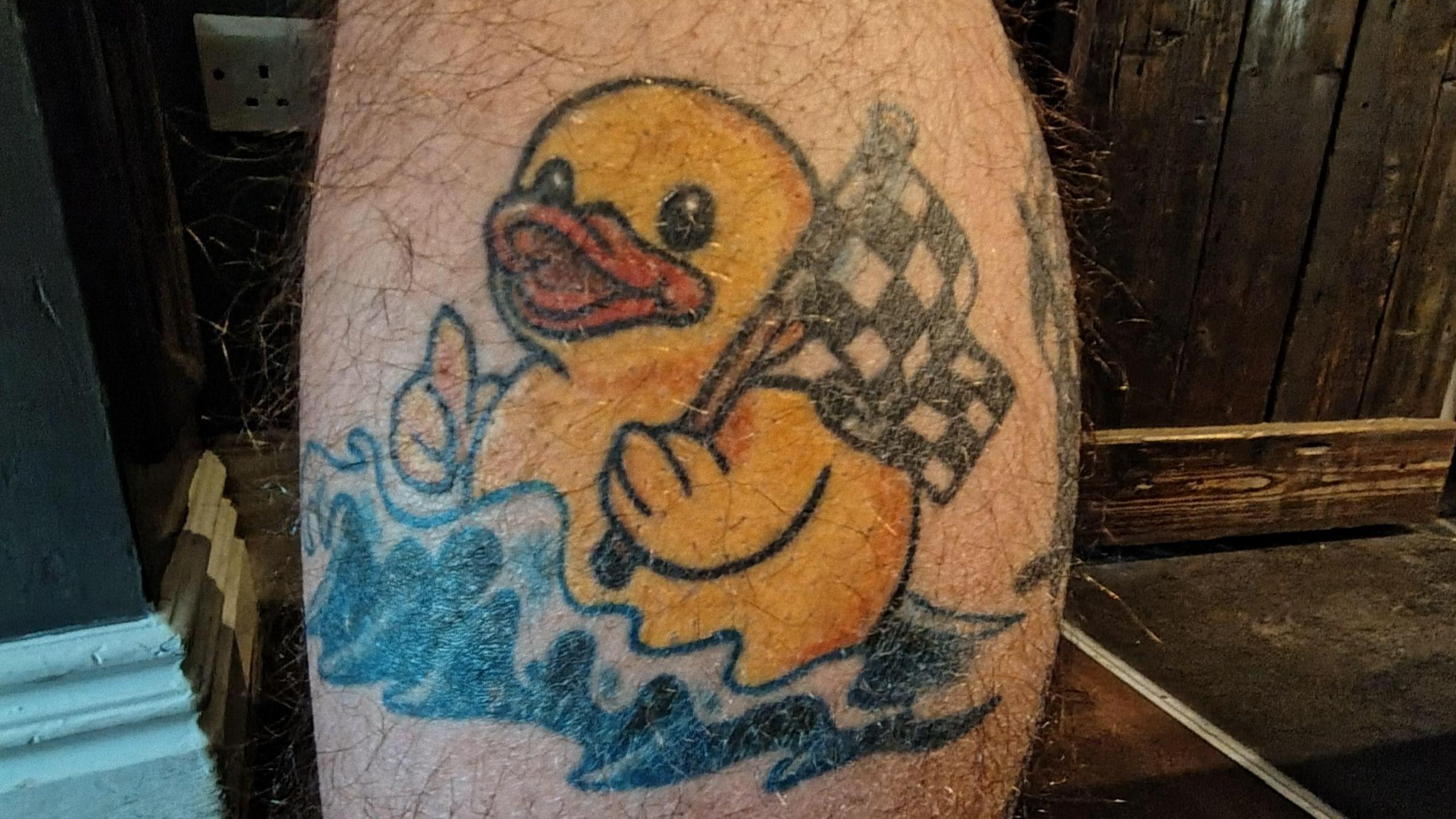 A close up of Curtis' leg. He has a yellow rubber duck tattoo. It has its thumb up and is holding a chequered flag. It is also in water.