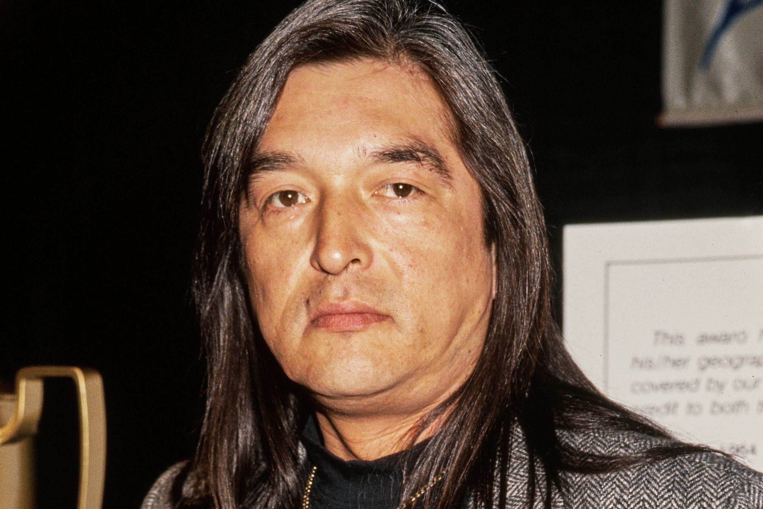 Graham Greene ka flokë të gjatë të drejtë dhe ka veshur një xhaketë me motive mbi një bluzë të errët.