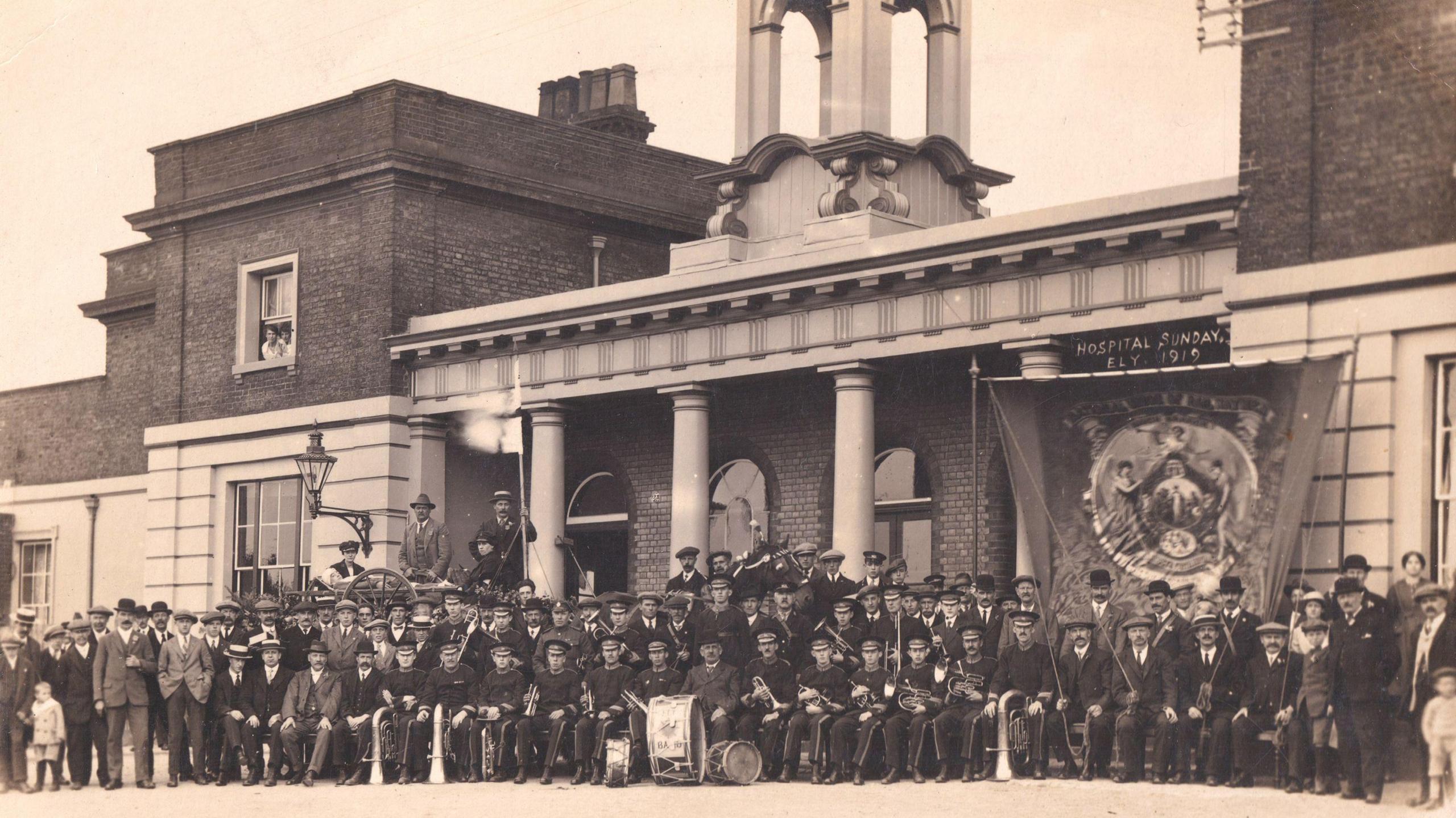 Black and white sepia photo from 1919 showing a large gathering of people outside Ely railway station, some form a brass band with their instruments around them.