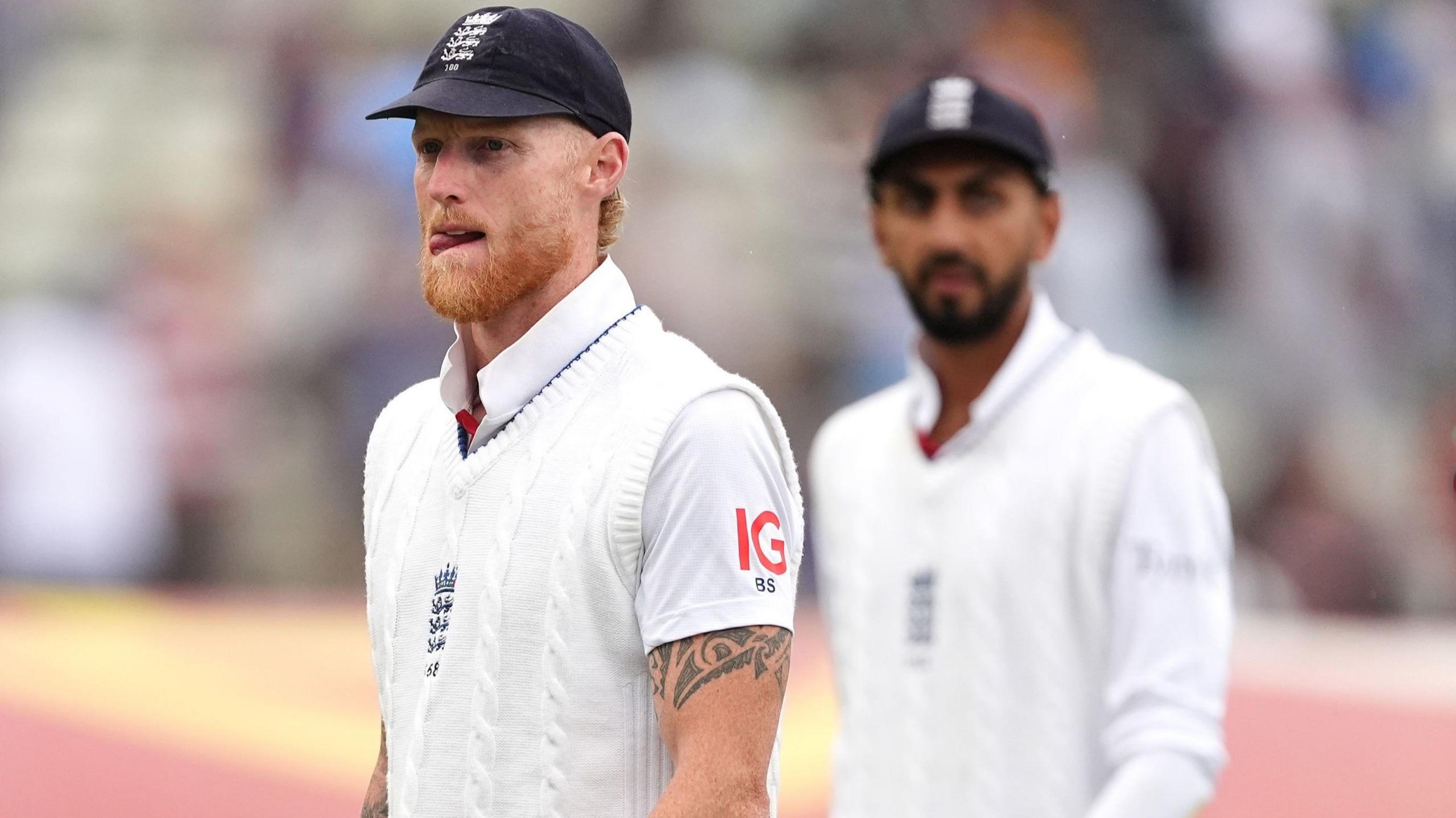 Ben Stokes, wearing a blue cap and white sweater, looks frustrated, left, with Shoaib Bashir in the background