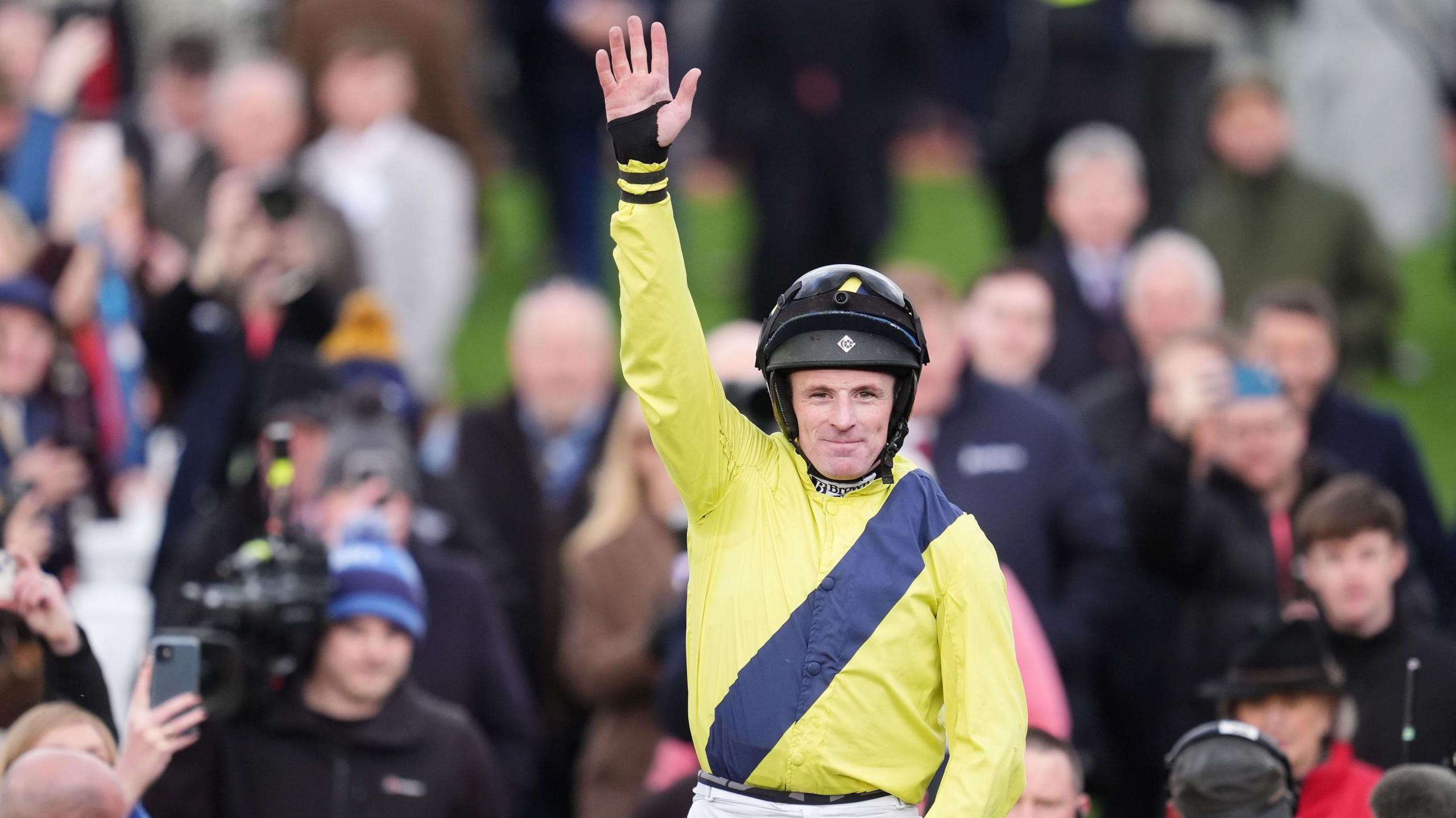 Jockey Sean Flanagan, dressed in yellow silks with a navy stripe across it rides his horse Marine Nationale to victory in the BetMGM Queen Mother Champion Chase. He's celebrating his victory with his arm in the air surrounded by racing fans cheering him on. 