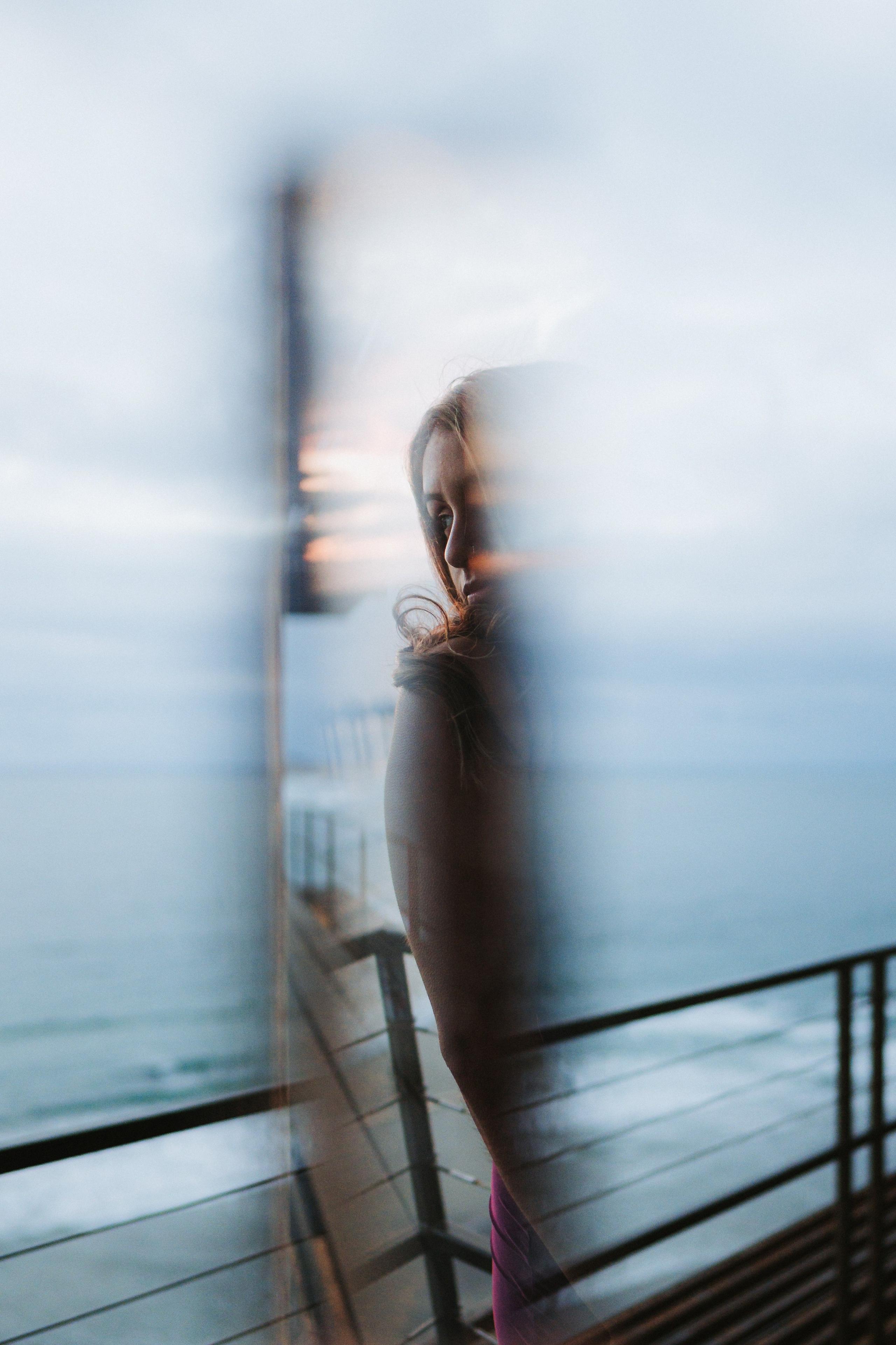 A woman photographed through glass. The image is blurred around the woman but she can be partly seen through the glass. She is looking over her shoulder and appears to be standing on a promenade beside the sea.