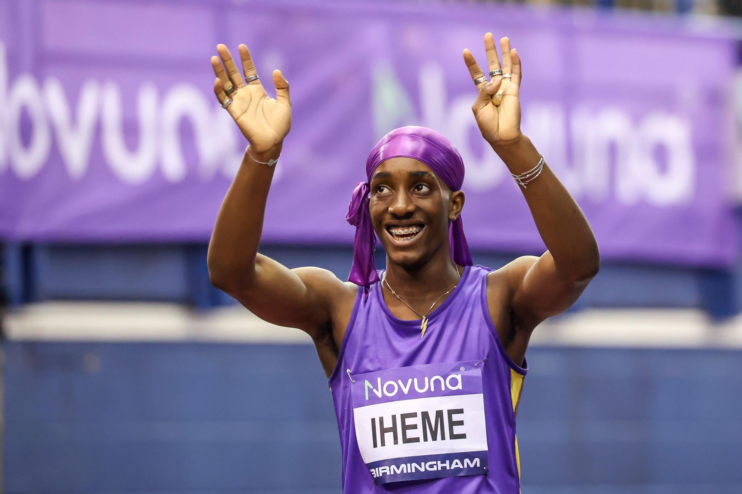 Divine Iheme wears a purple durag and a purple tank top with his name on a bib on the front. He is holding his arms up in the air and waving at the crowd. He has a big smile on his face and you can see his braces. He is wearing a gold necklace with a lightning charm on it.