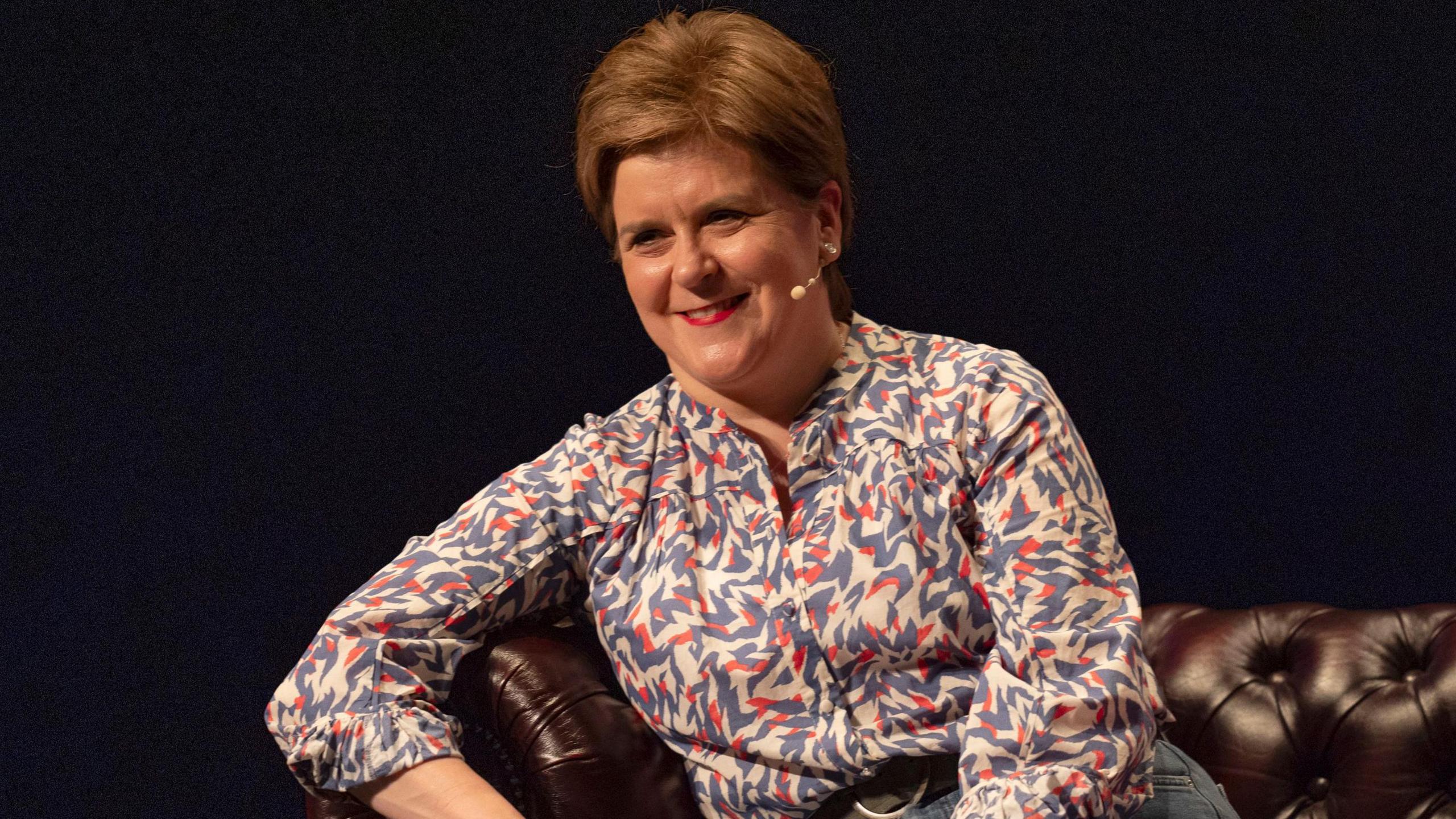 Nicola Sturgeon is sat on a brown leather sofa.
She has a lip mic on.
She is wearing a pale blue, white and red blouse and has short reddish hair.