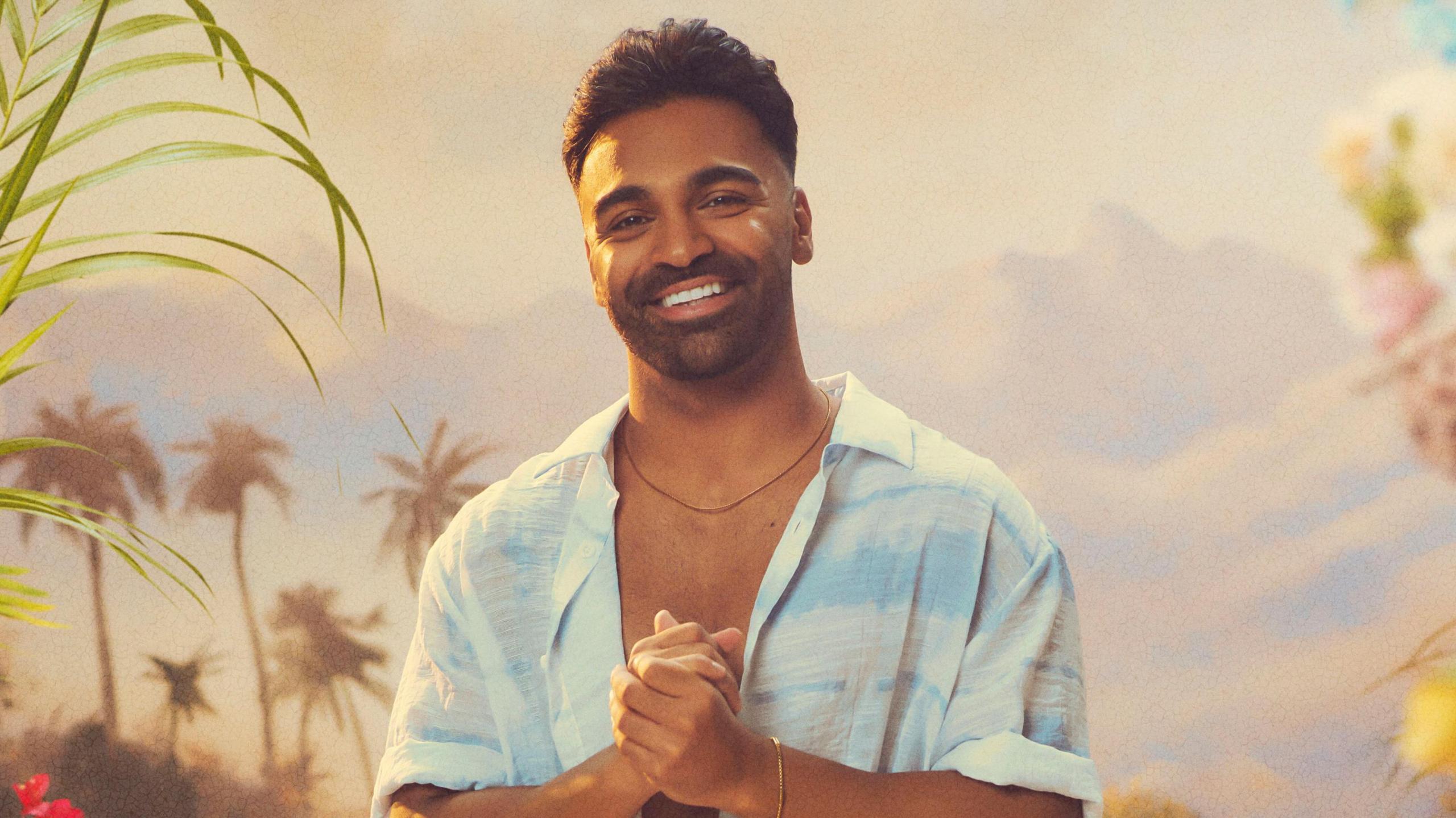 Nas Majeed poses with his hands clasped for a promo shot for the new series of All Stars. Nas has curly brown hair and a trimmed beard and wears an unbuttoned blue tie-dyed shirt.