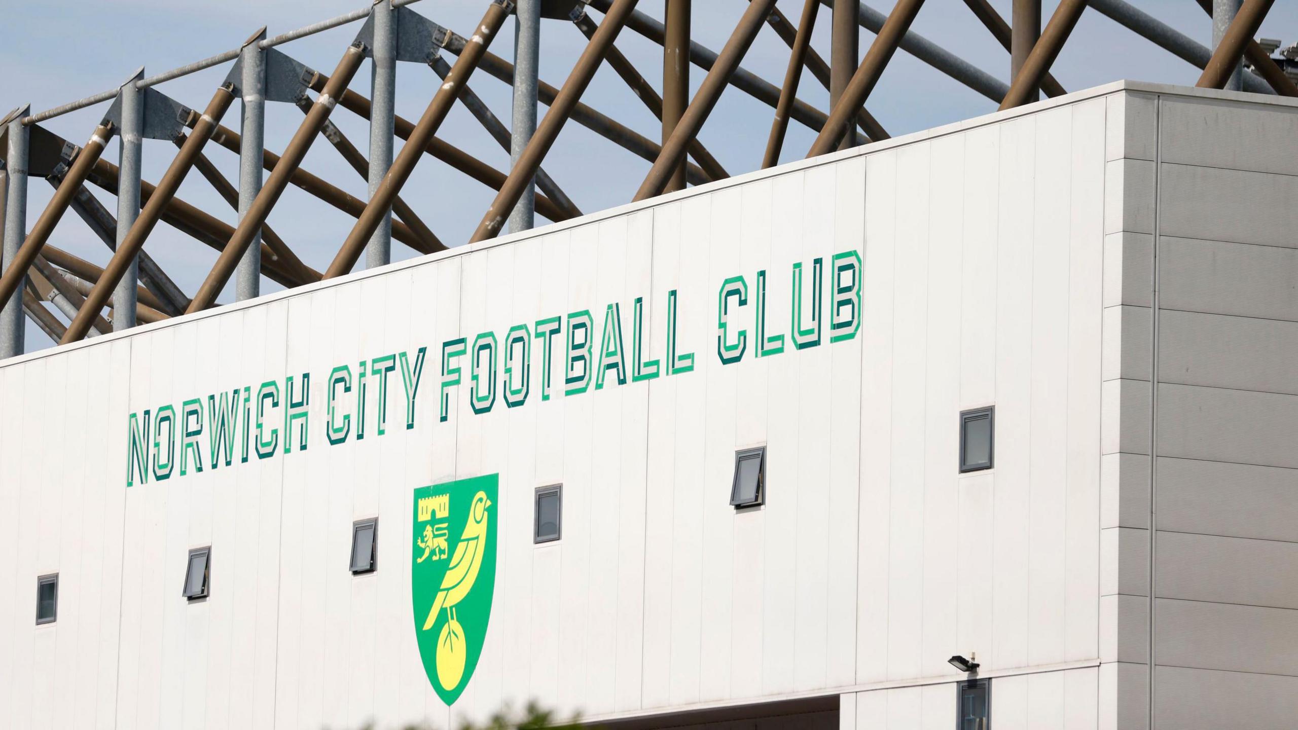 A close up of the Norwich City Football Club sign and logo which are painted on an outside wall of the club's Carrow Road stadium. The writing is in green and the logo is a shield shape with a yellow canary standing on a yellow ball. The background of the shield is green.