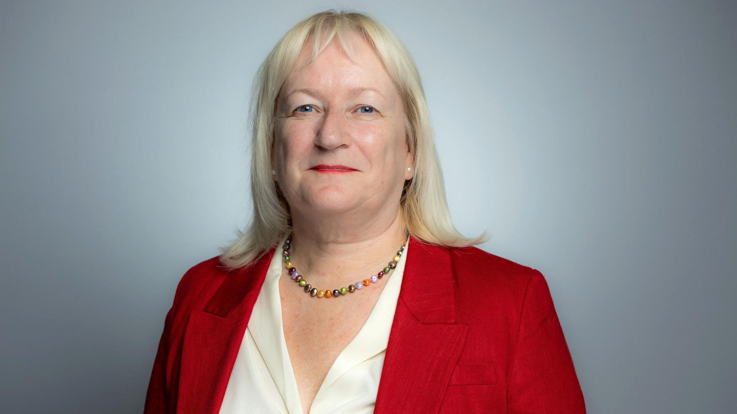 A blonde woman wearing a red blazer, a white shirt and a rainbow beaded necklace. She's looking straight at the camera and smiling against a grey wall.