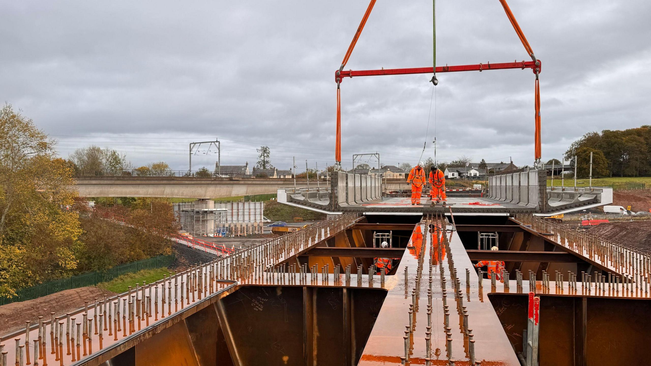 The steel structure which will hold the deck of the Clifton Bridge near Penrith. Two people in orange high viz clothing and wearing a safety harness look down below the deck, where two other workers are. The old concrete is in the distance with the roofs of houses visible behind.