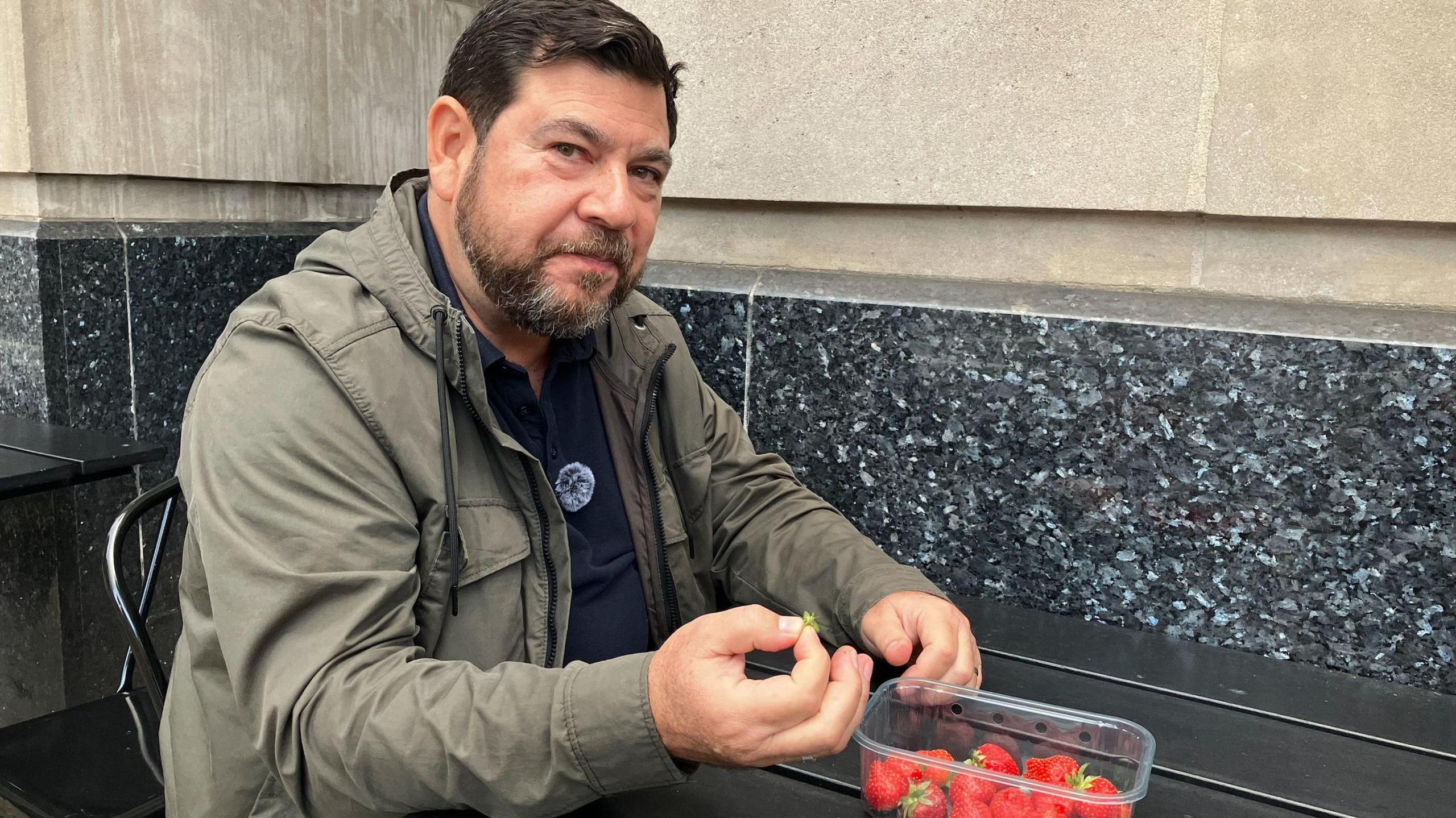 Kleo Papas with dark hair and a beard wearing a khaki jacket sits at an outdoor table holding a strawberry stalk beside a plastic punnet of strawberries. A stone building wall is behind him.