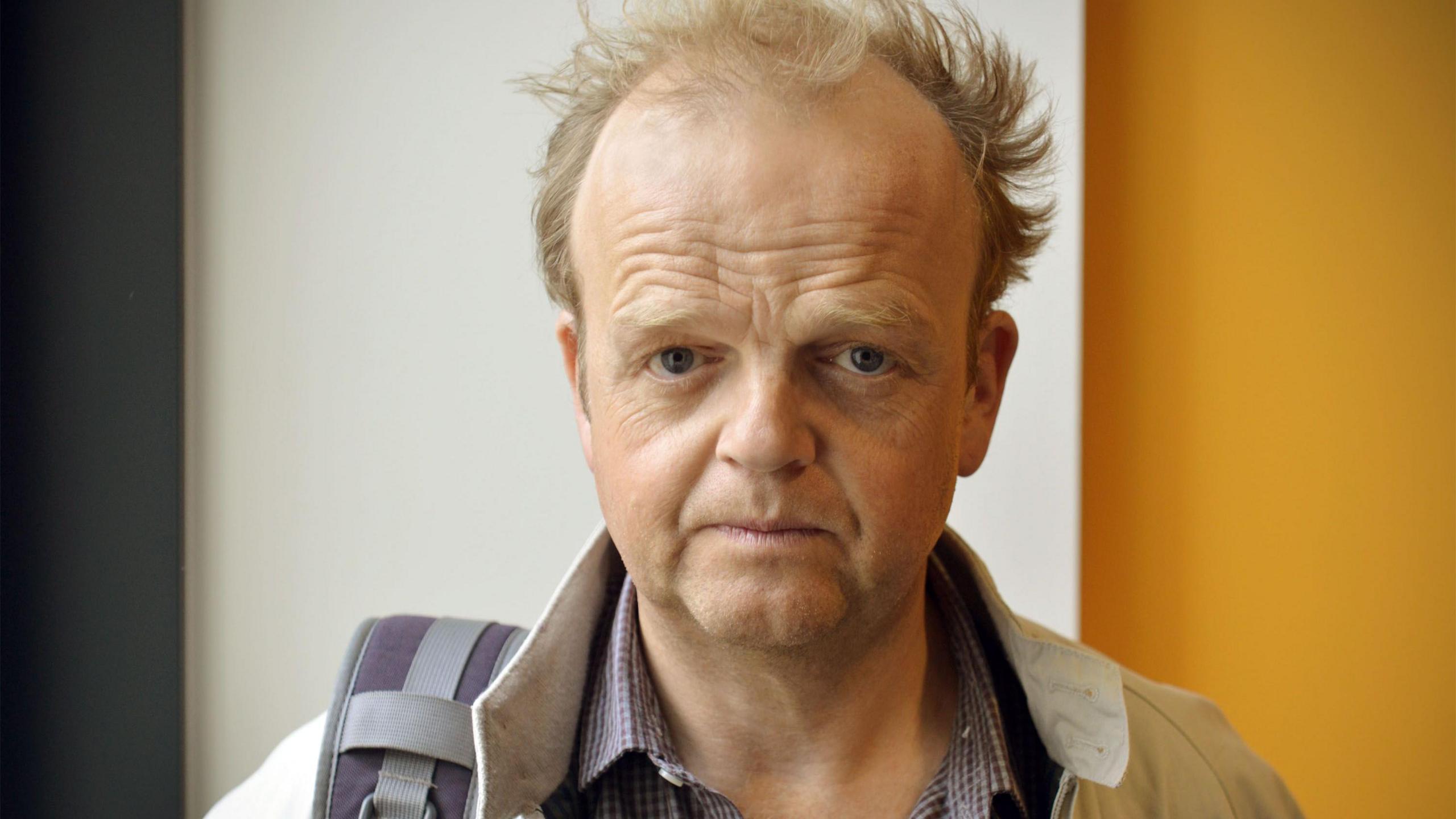 Actor Toby Jones. A middle-aged man with a receding hairline and reddish brown hair looks at the camera with a serious face. He is  wearing a purple checked shirt and a tan jacket and has one backpack strap over his shoulder.