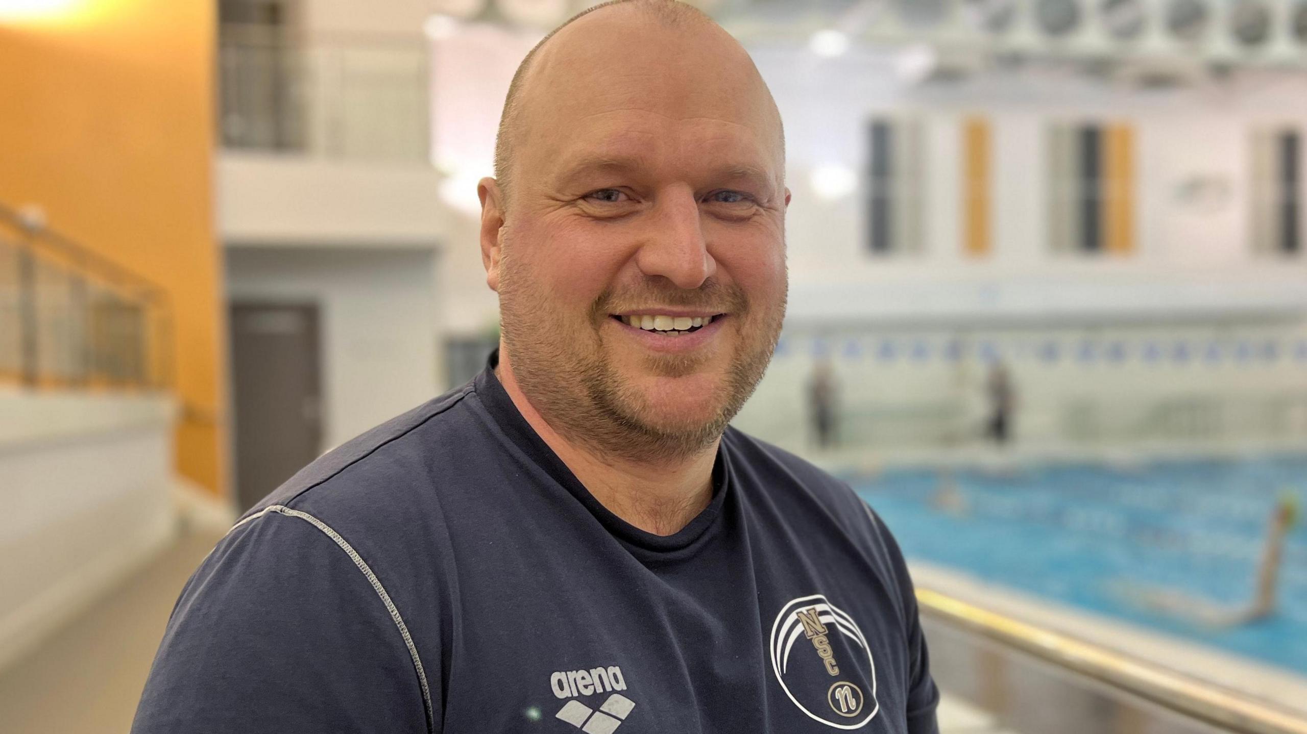 Andy Sharp wearing a navy Northampton Swimming Club. He is looking directly at the camera with a broad smile and people can be seen swimming in a blurred background