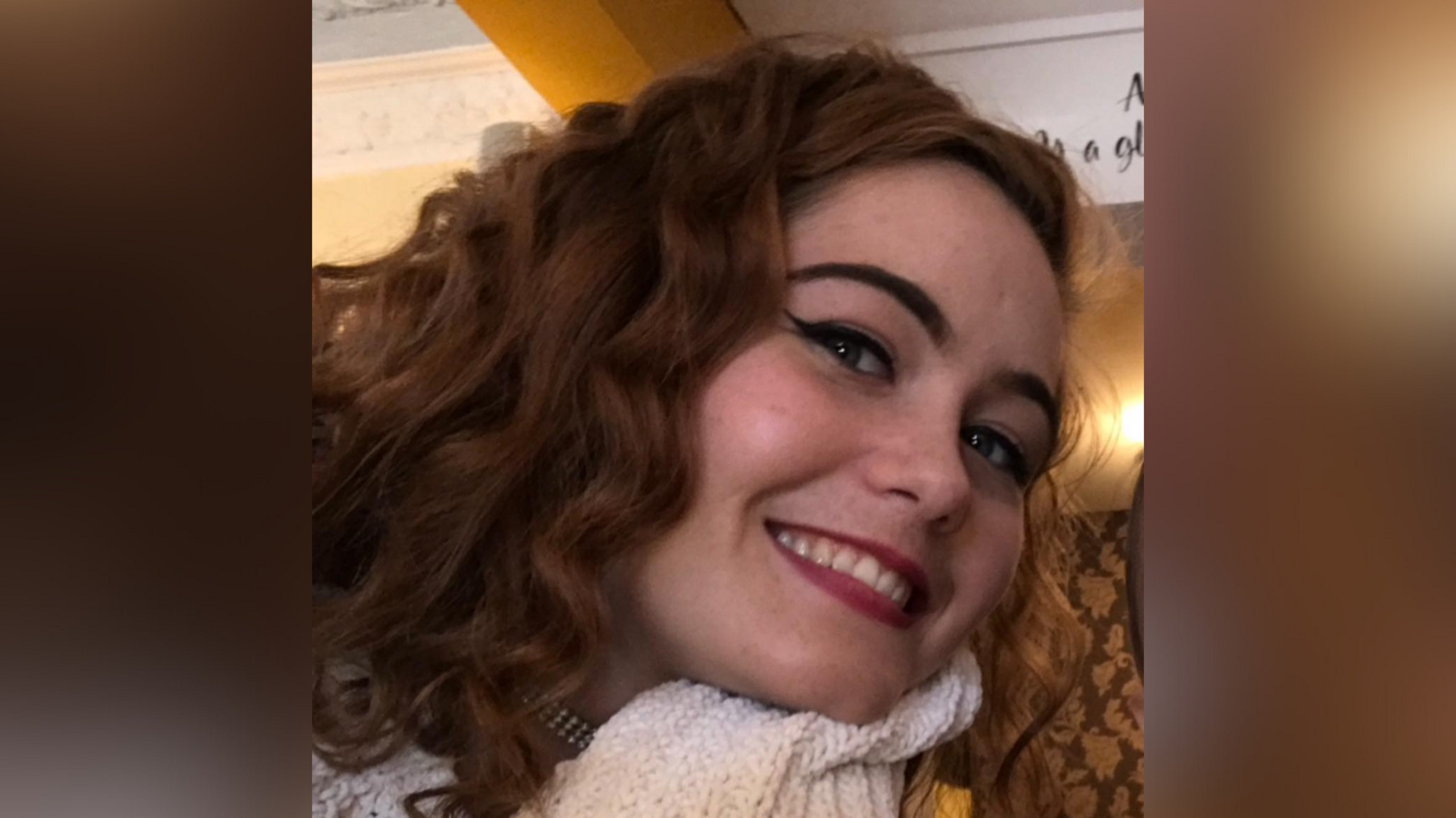 A smiling Courtney Mitchell looks directly at the camera with her head tilted to her left. She is a young woman with long, slightly curly dark brown/ginger hair and is wearing a white scarf.