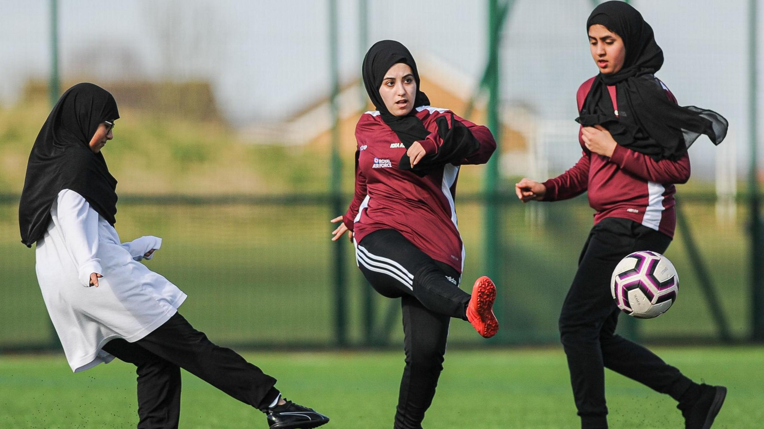 Three girls kick a football between themselves on a pitch. Two of the girls are wearing black head scarves, red shirts and black trousers and one is wearing a white shirt. 