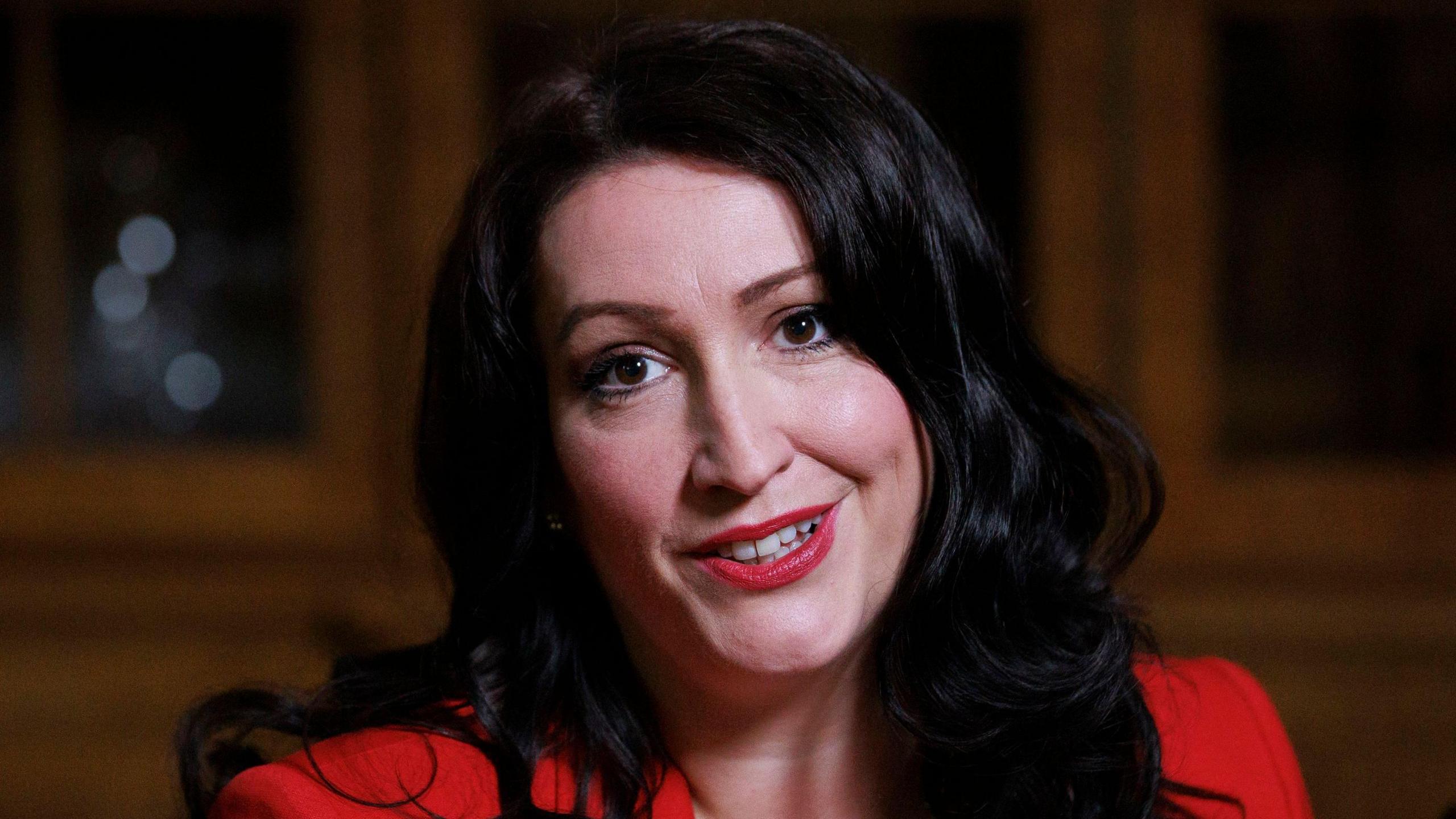 Deputy First Minister Emma Little-Pengelly wearing red with red lipstick and black curly hair smiling at the camera. 