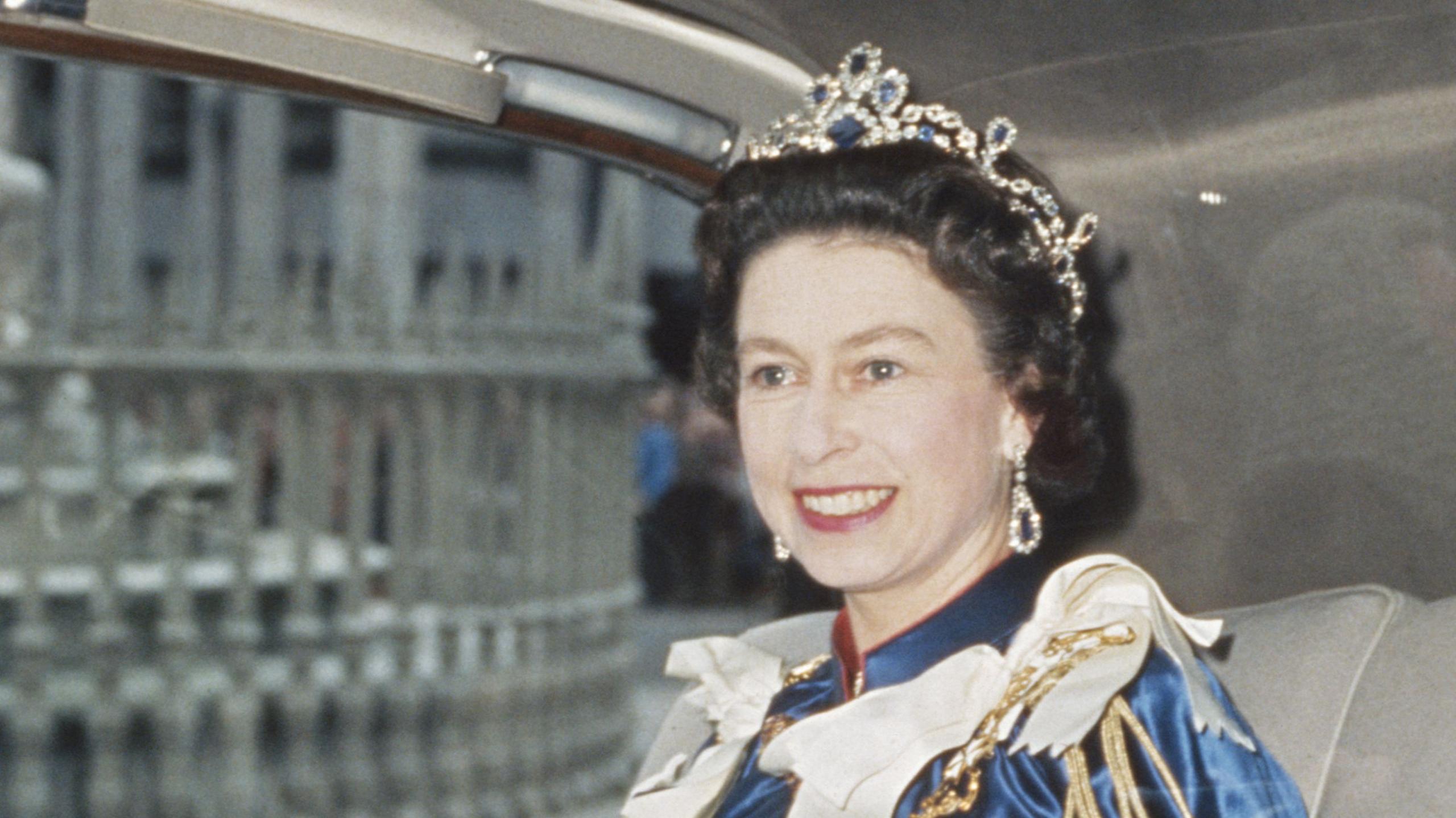 Queen Elizabeth, in royal regalia including a crown, smiling and riding in a car in the late 1960s.