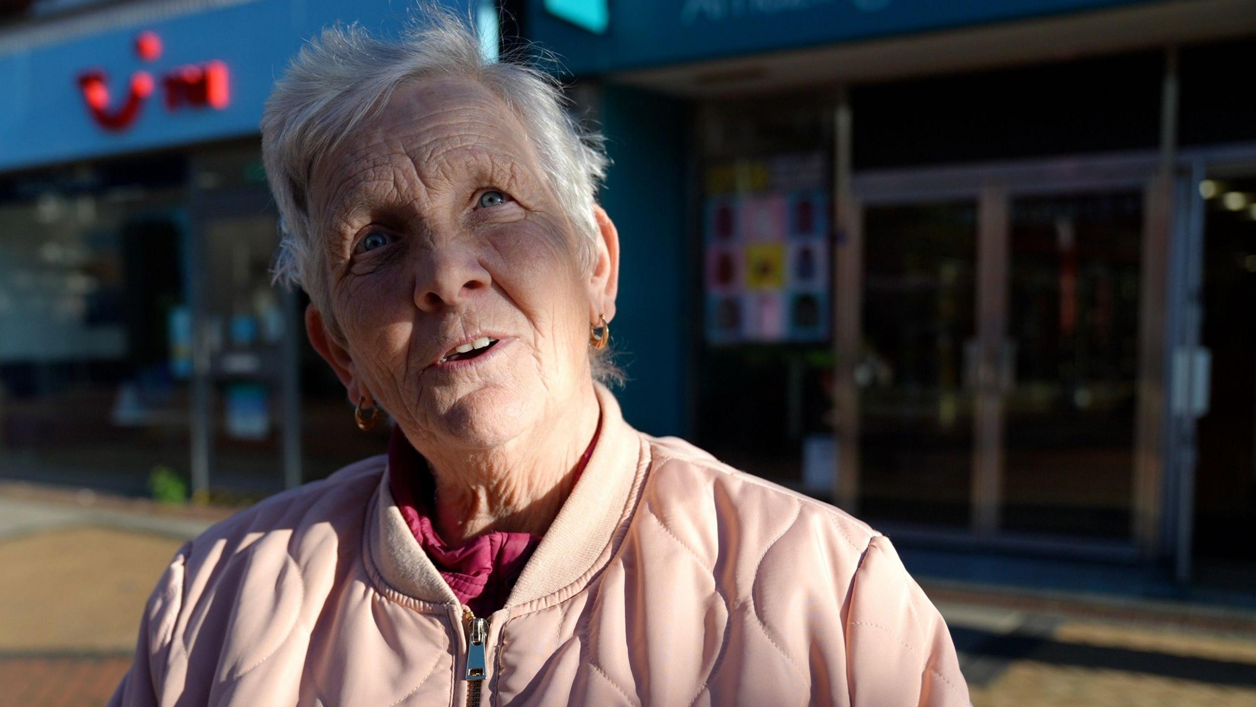 A woman with short white hair looks up as if in thought. She is standing on a high street and wearing a light pink jacket.