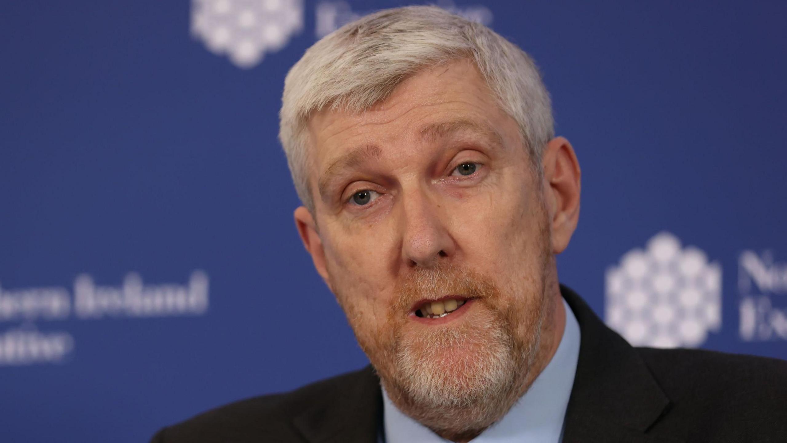 John O'Dowd who has short white hair and a beard. He is speaking at a conference, wearing a black suit and blue shirt.