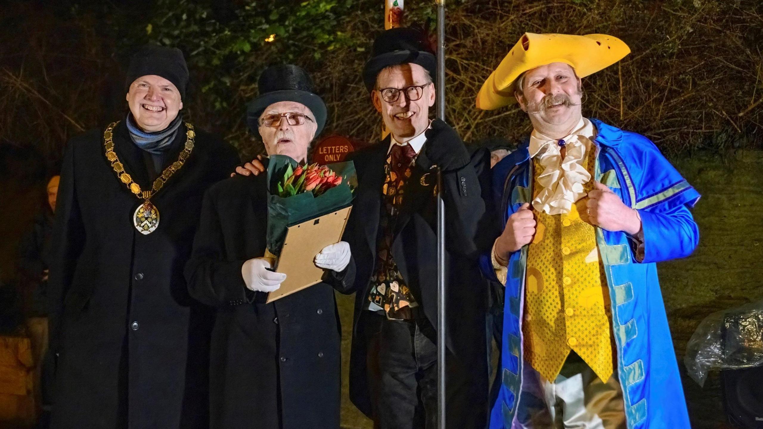 Four men stand together under a lamp in Frome. Three of them are dressed in traditional dark outfits, with two wearing black top hats, while the fourth is wearing a bright yellow waistcoat, a bright blue long jacket and a yellow pirate-style three cornered hat