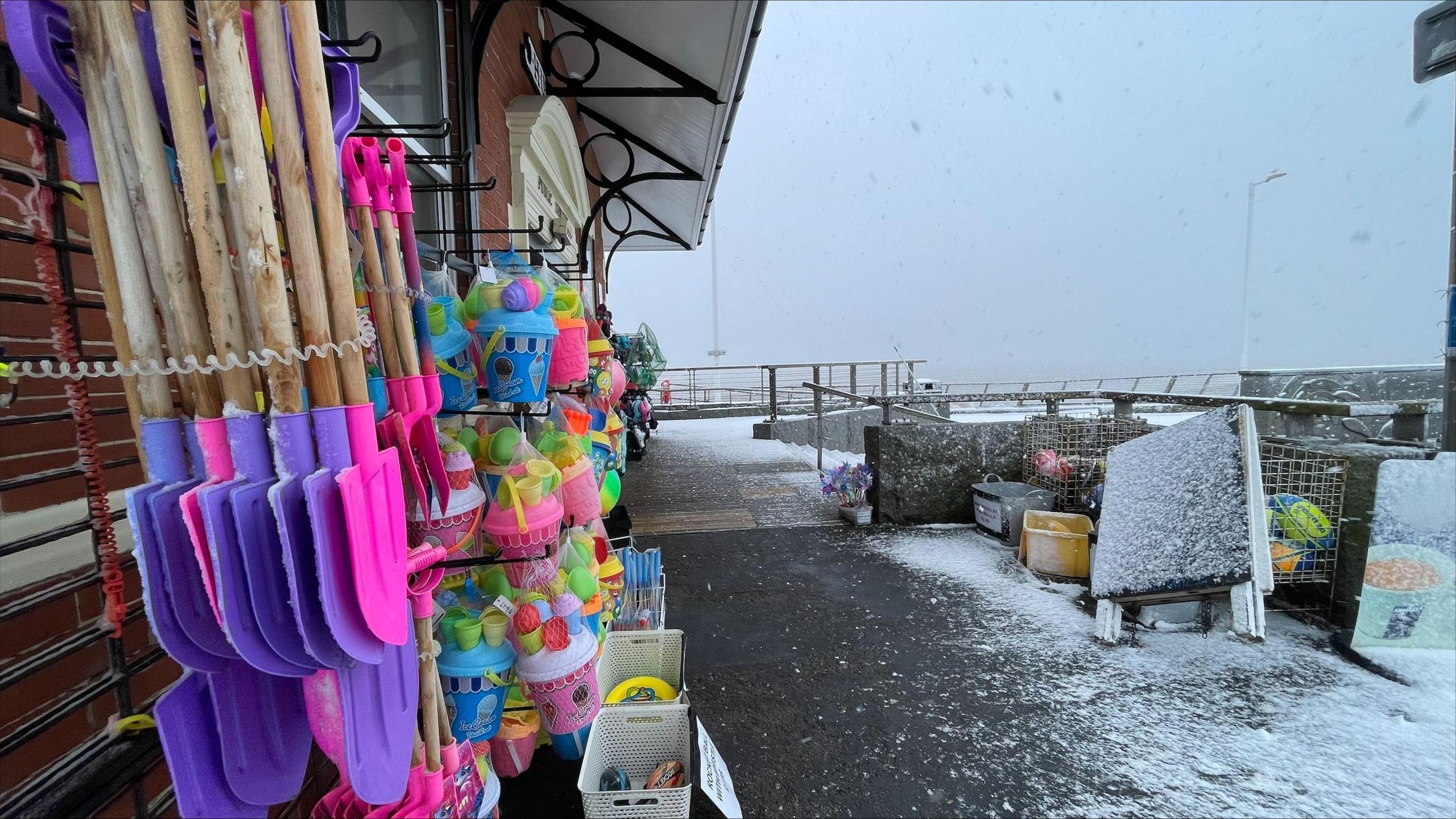 Pink and purple toy beach spades hanging outside a shop. Next to it is a row of buckets and toys