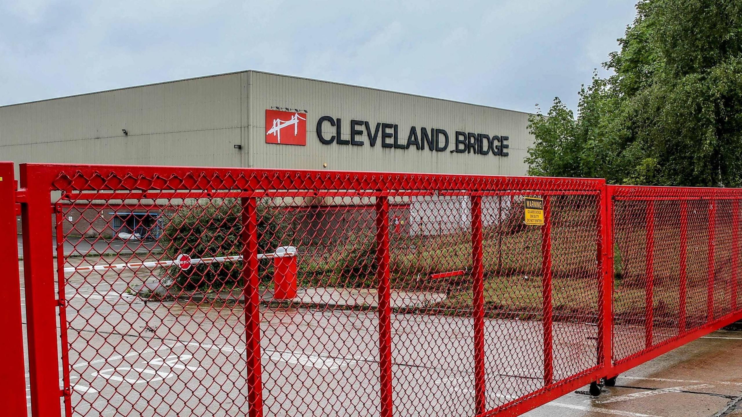 The outside of the Cleveland Bridge site with a large red metal gate which is closed. 'Cleveland Bridge' is written on the side of a large grey factory.