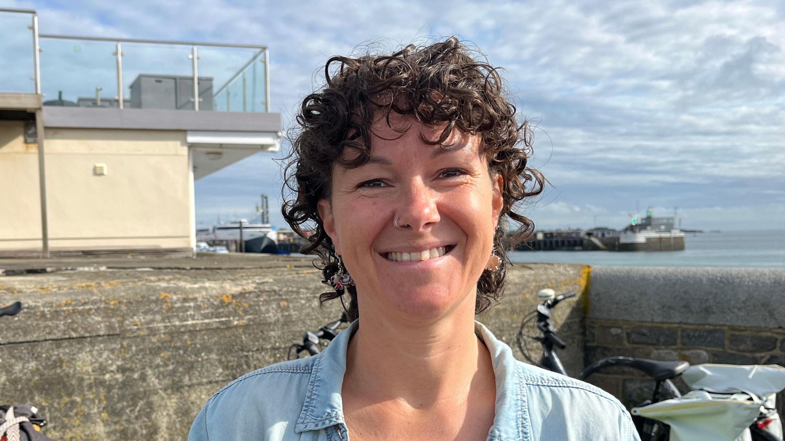 Tina Bury is standing outside in front of a concrete wall. A number of push bikes are behind her. She is wearing a blue shirt and is smiling into the camera. The sea is in the background.