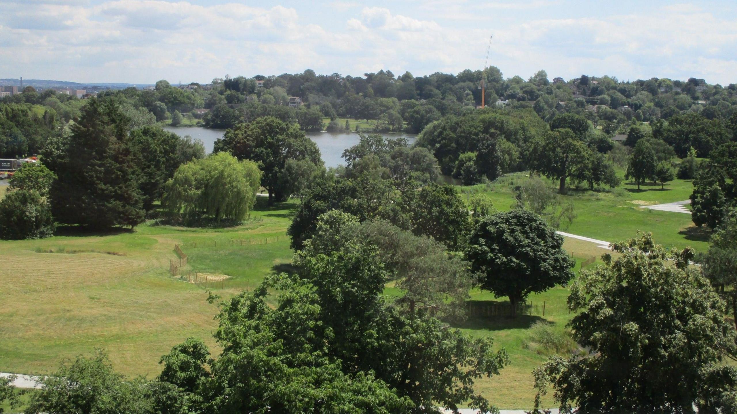 An aerial view of the current golf course within Wimbledon Park on a sunny day with blue skies. There are a large number of trees in the shot and a lake in the middle.