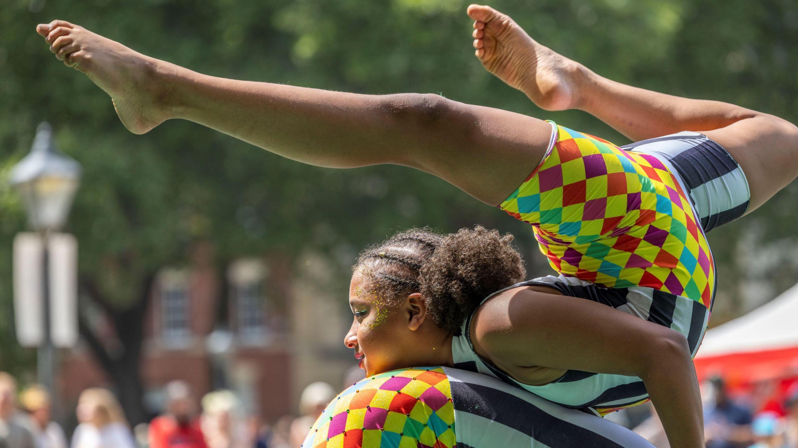 A young person in a colourful leotard doing a gymnastic trick on the back of someone else in the same outfit