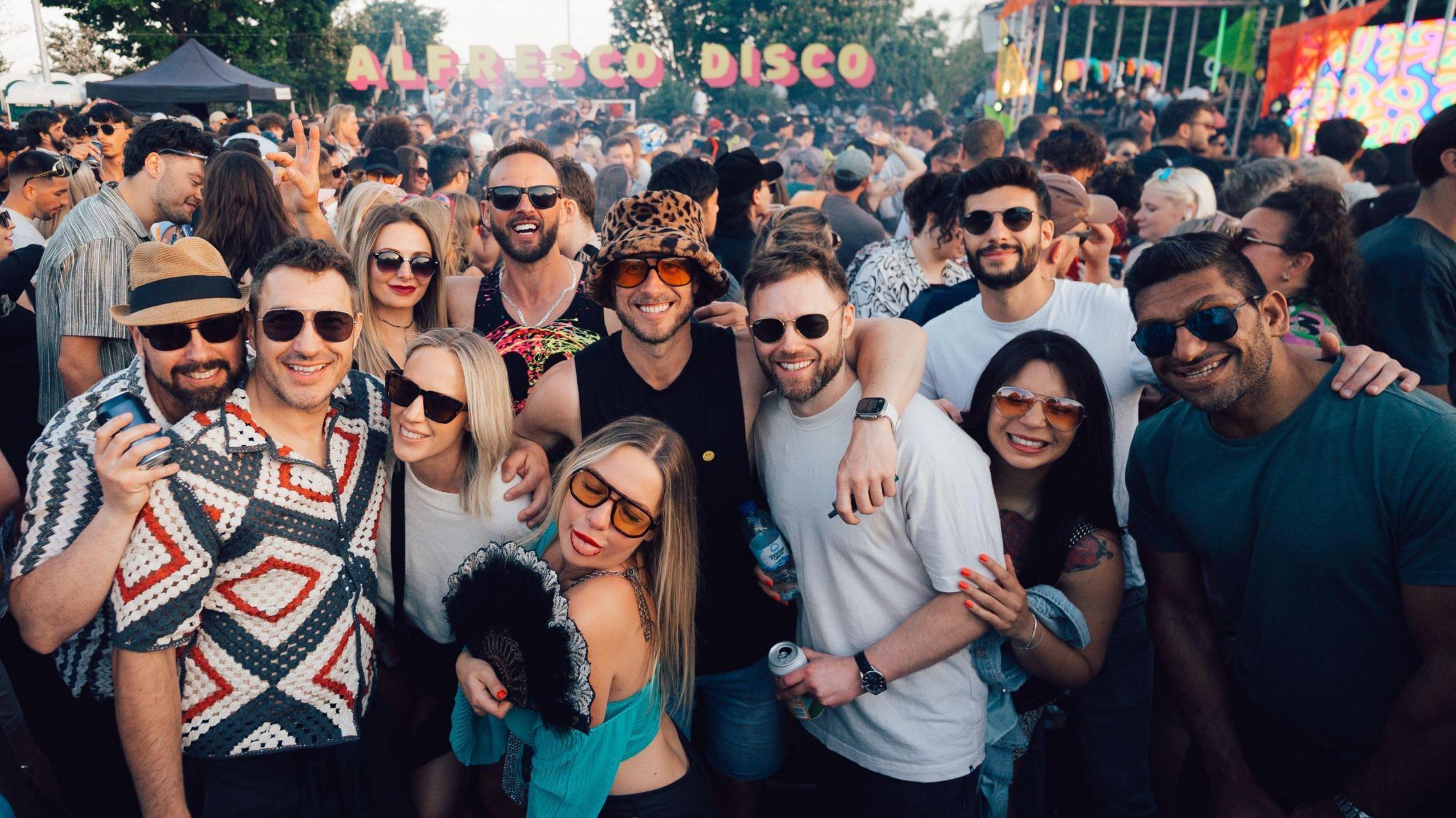 A large group of young people smile at the camera as they pose in a group shot at Alfresco Disco in Bristol. In the background is a large crowd dancing to the music