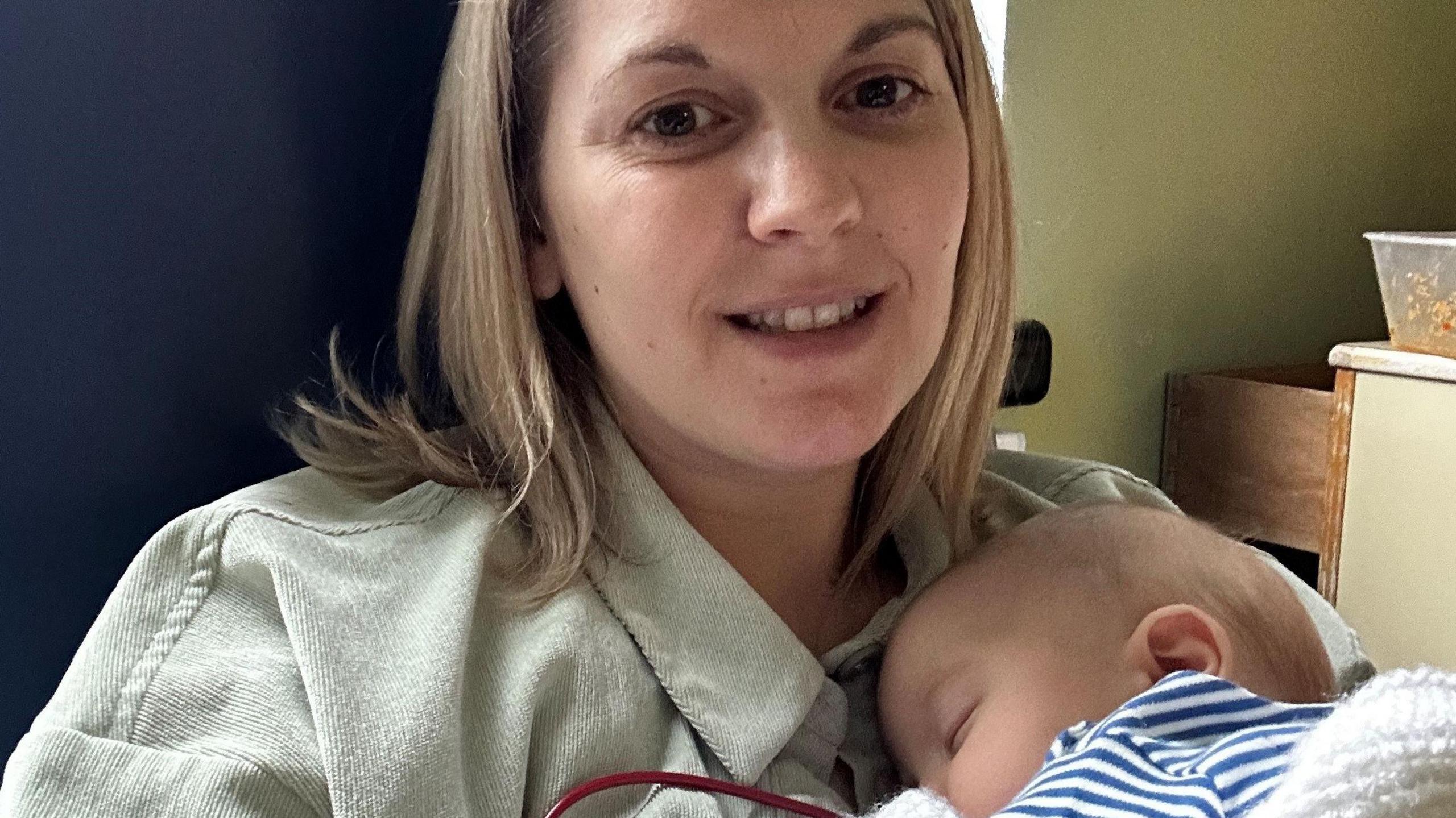 Polly sits in a chair, holding a sleeping baby. She has shoulder length dark blonde hair and is smiling at the camera.