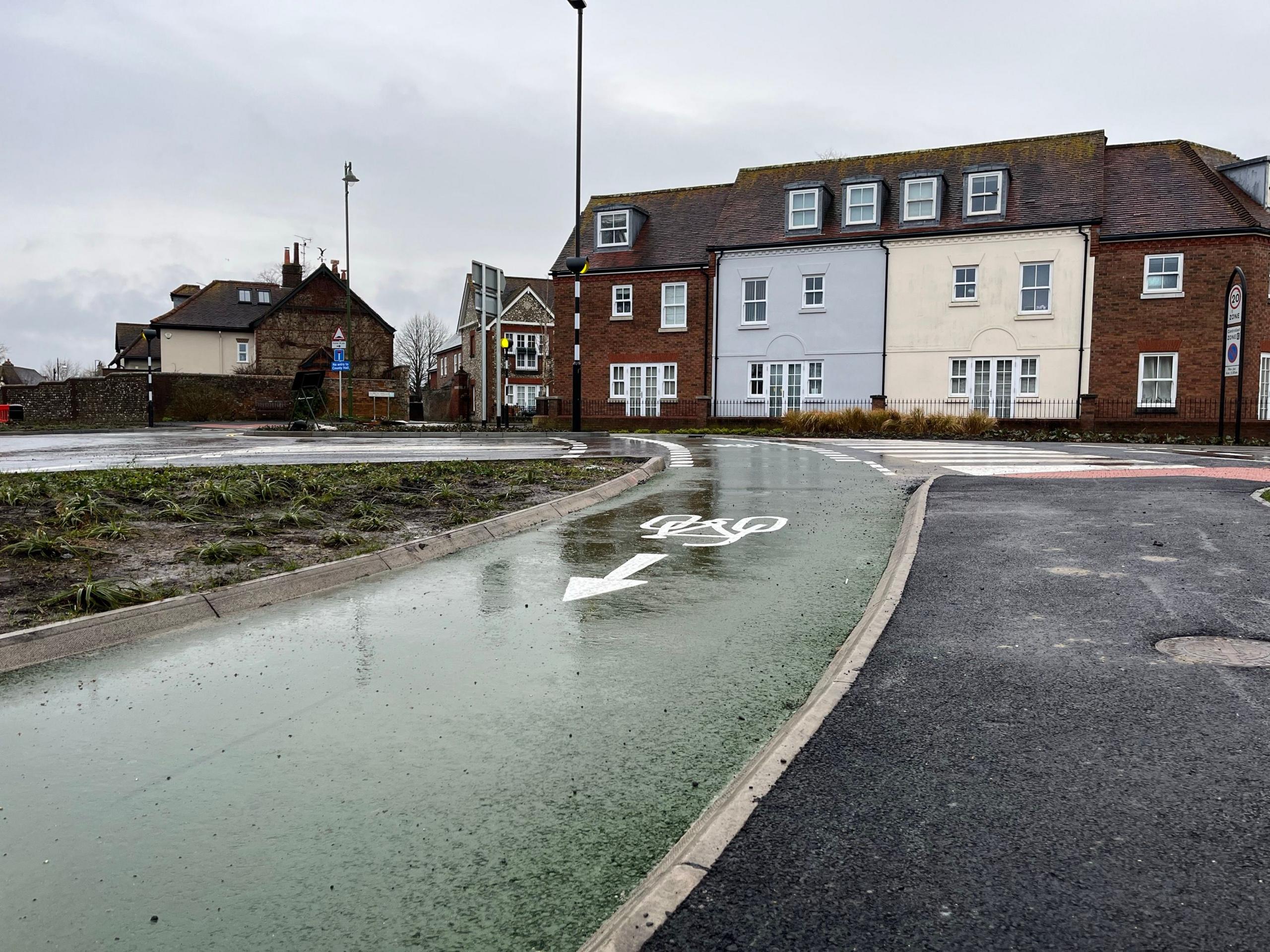 The green cycle lane which goes around the roundabout's perimeter. It's reflecting the sky as it has been raining. There is an icon of a bike and arrow painted on the lane in white. In the background are houses and one of the crossing points