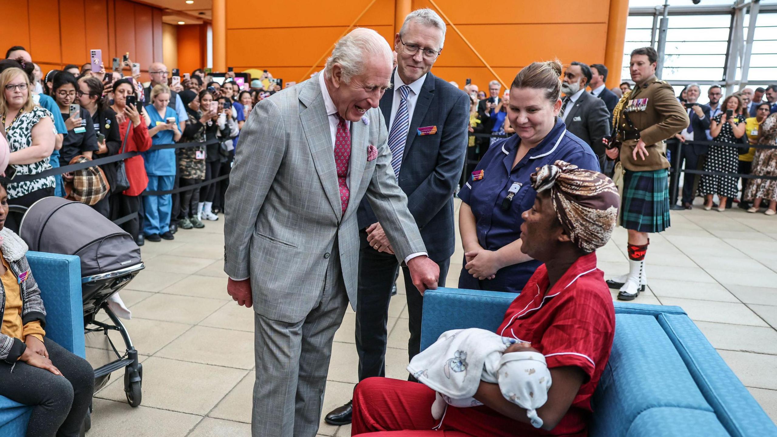 The King chats to a woman sitting on a blue sofa, wearing red pyjamas and a brown headscarf. She is holding an infant who has a hat and a blanket draped across her. The King is beaming as he leans down to chat to the woman, who is also smiling. A man in a suit and a nurse smile as they look on, while crowds of hospital staff stand behind black tape.