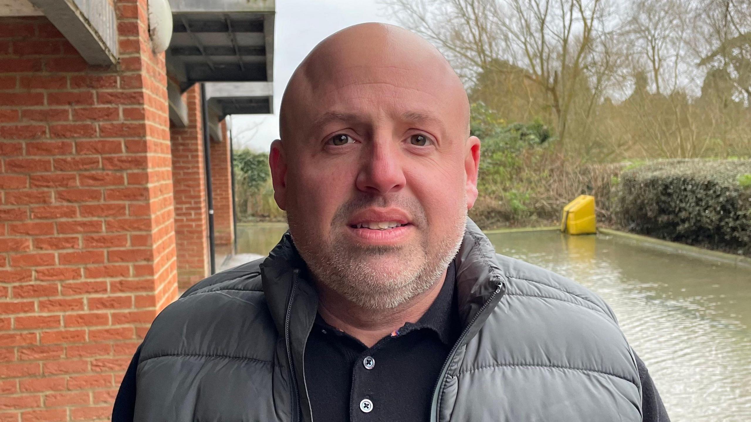 Richard Ayres looking direct to camera, standing outside in the car park belonging to his block of flats. He has a bald head and brown eyes, and is wearing a black collared T-shirt and grey gilet. To the right of the image is water covering part of the car park. A yellow grit bin can be seen in the corner, surrounded by water. To the left of the image is the brickwork of the apartment block. 