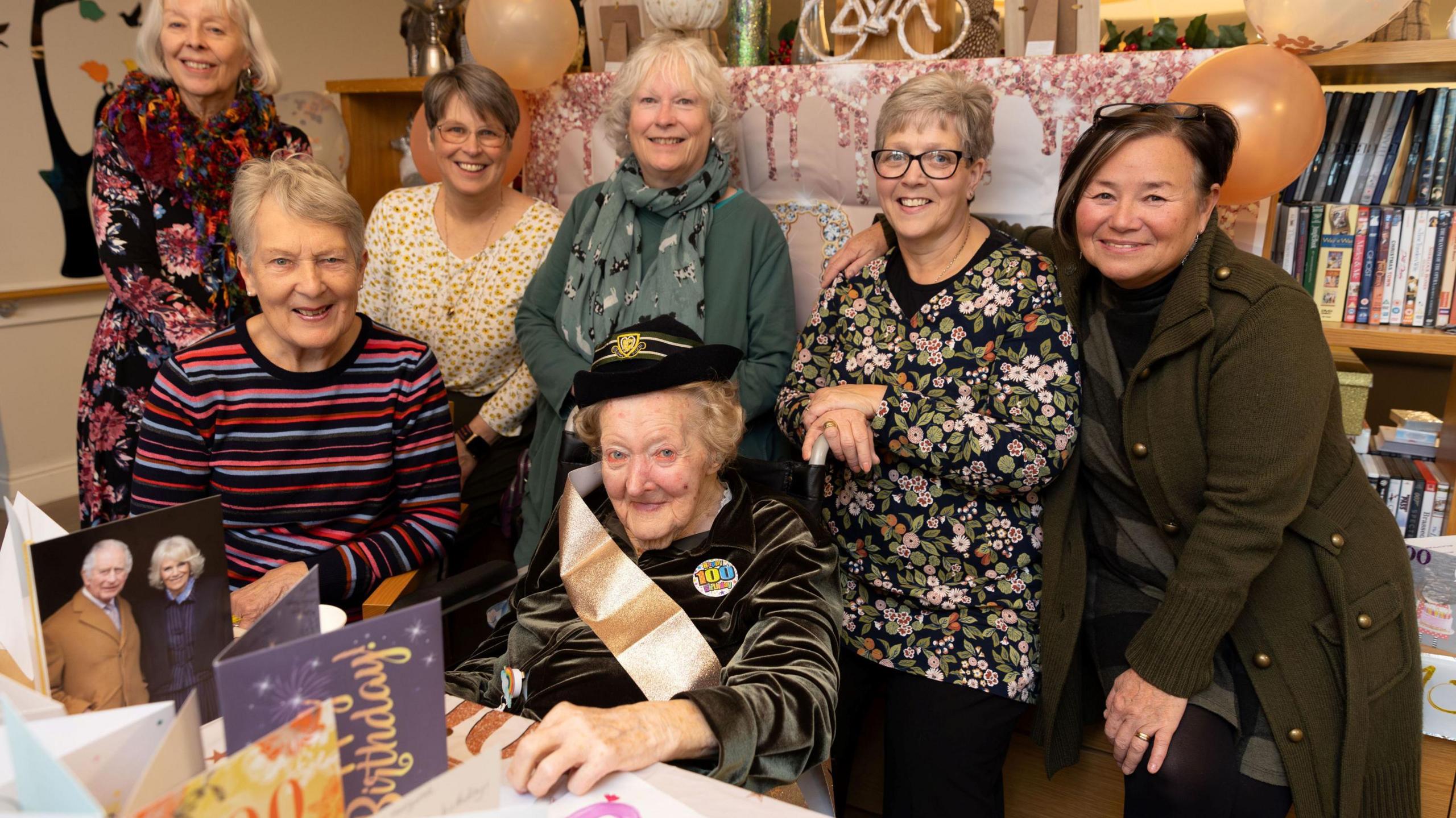 An elderly woman wearing a bright birthday sash sits at a table with several other women standing behind her in a group as they all smile at the camera. The picture is taken at St Monica Trust's Charterhouse Care Home in Keynsham and the woman's name is Margaret Grey.