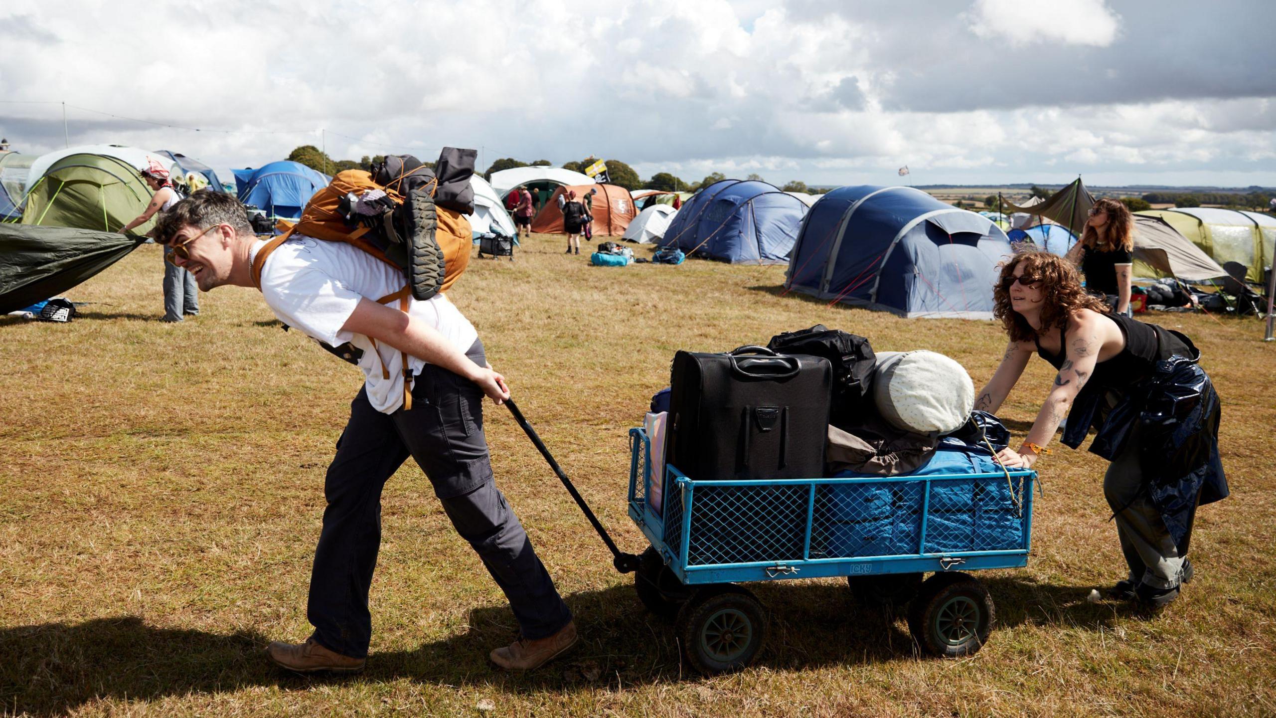 A couple propel their small trailer across the campsite at the End of the Road festival, with the young man pulling and his female partner pushing. In the background other tents can be seen set up