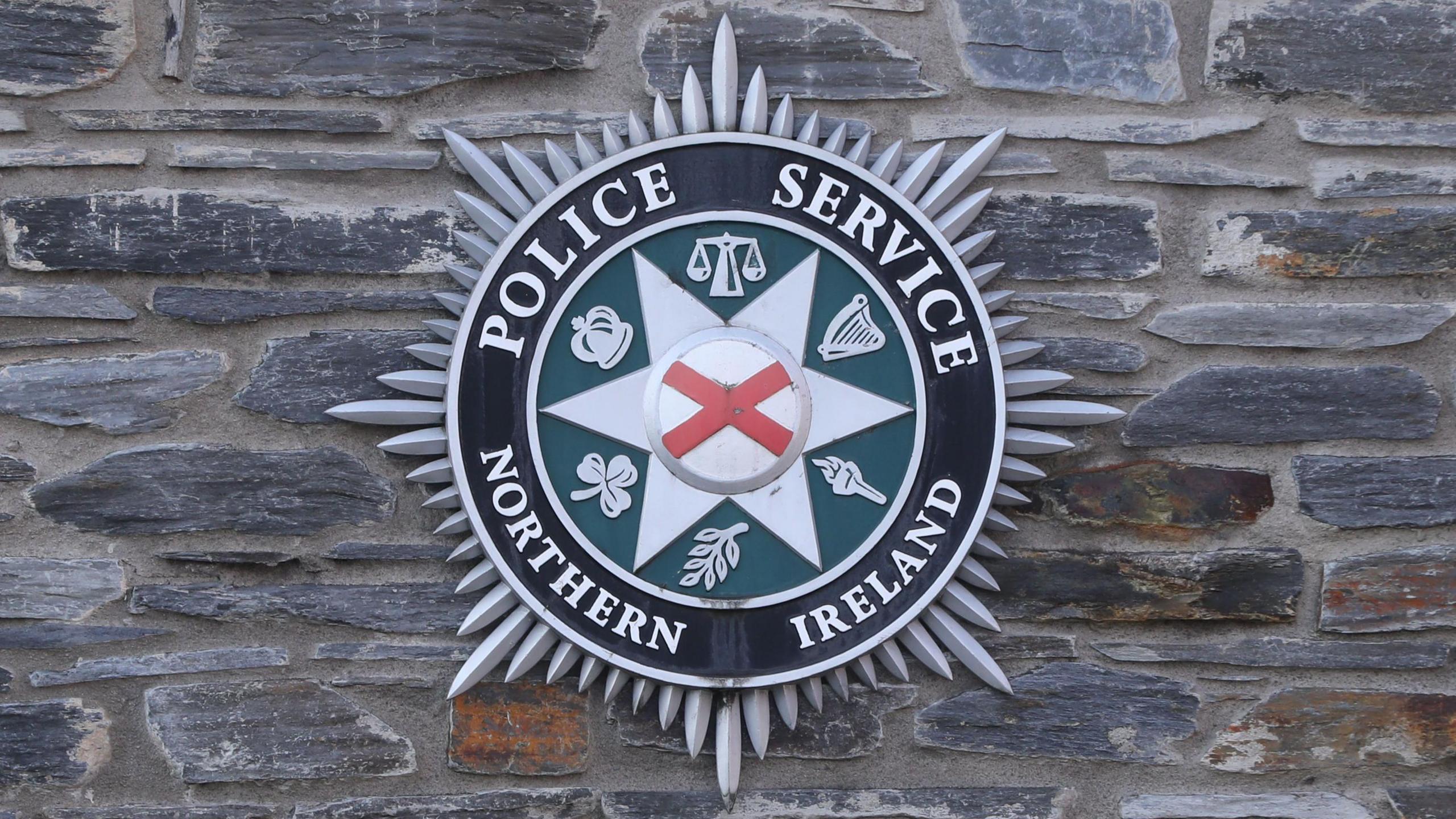 PSNI crest which says Police Service Northern Ireland - with emblems of a crown, balanced scales, harp, shamrock, torch and branch. The crest is silver, black and green with a white and red cross in the centre. 