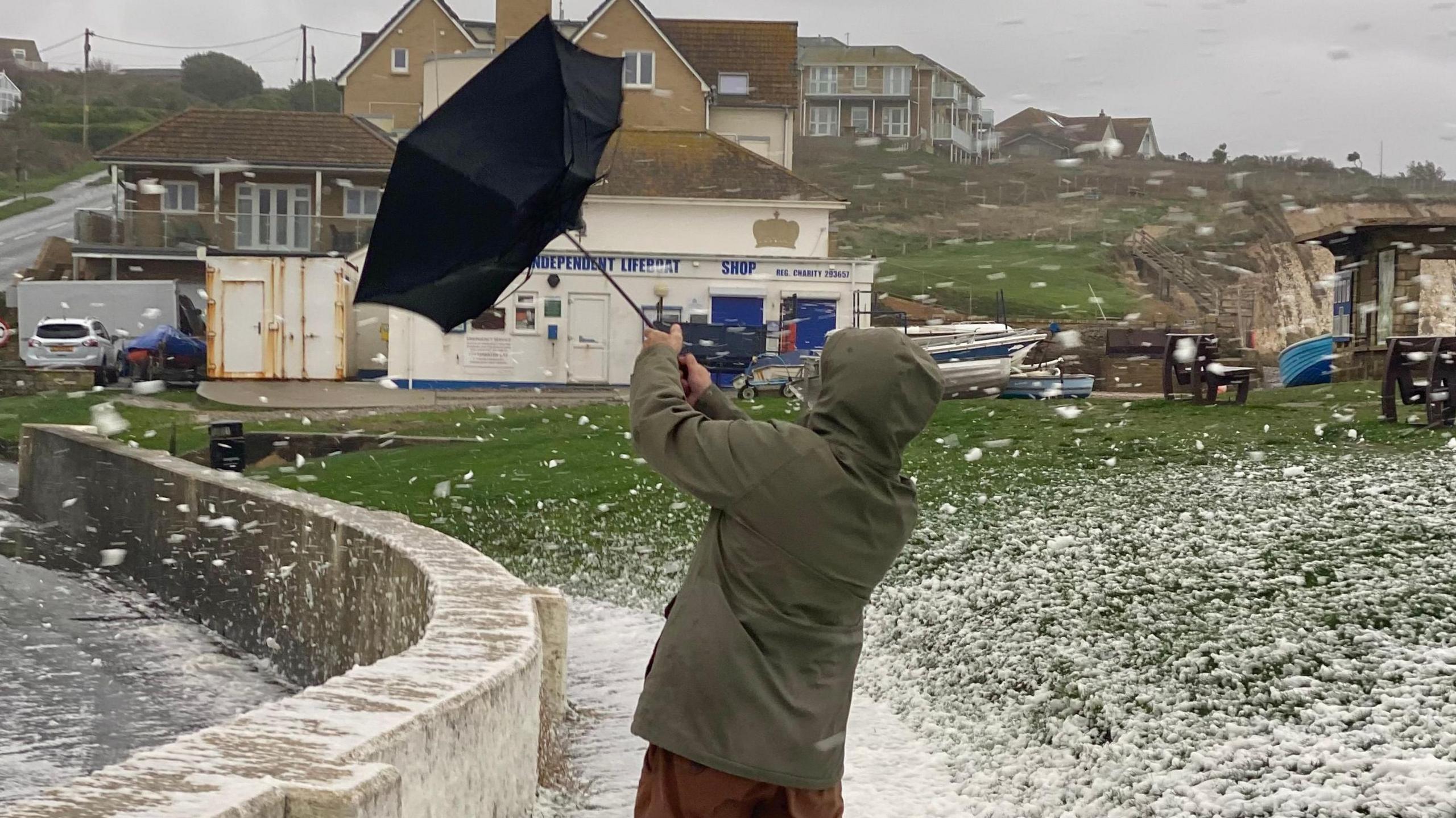 A person in a winter coat with a hood holds an umbrella which is blown inside out by the wind. Spray from the sea flies through the air and over a coastal wall onto a path.