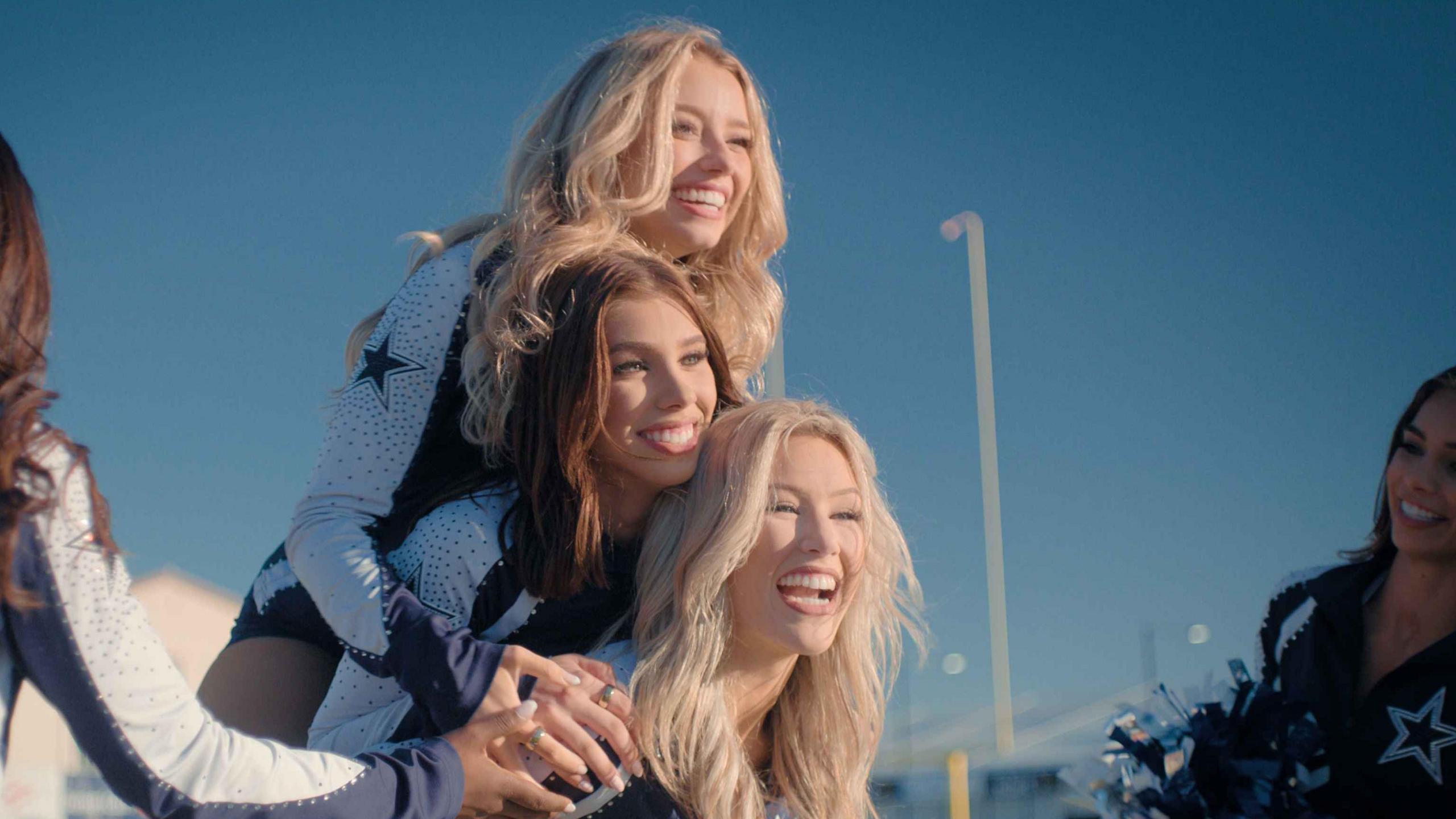 Pictured from top to bottom: Kleine, Lea, Megan. Kleine and Megan have long blonde hair and Lea has long brunette hair. They are shown jumping on each other while two squad members stand at the side
