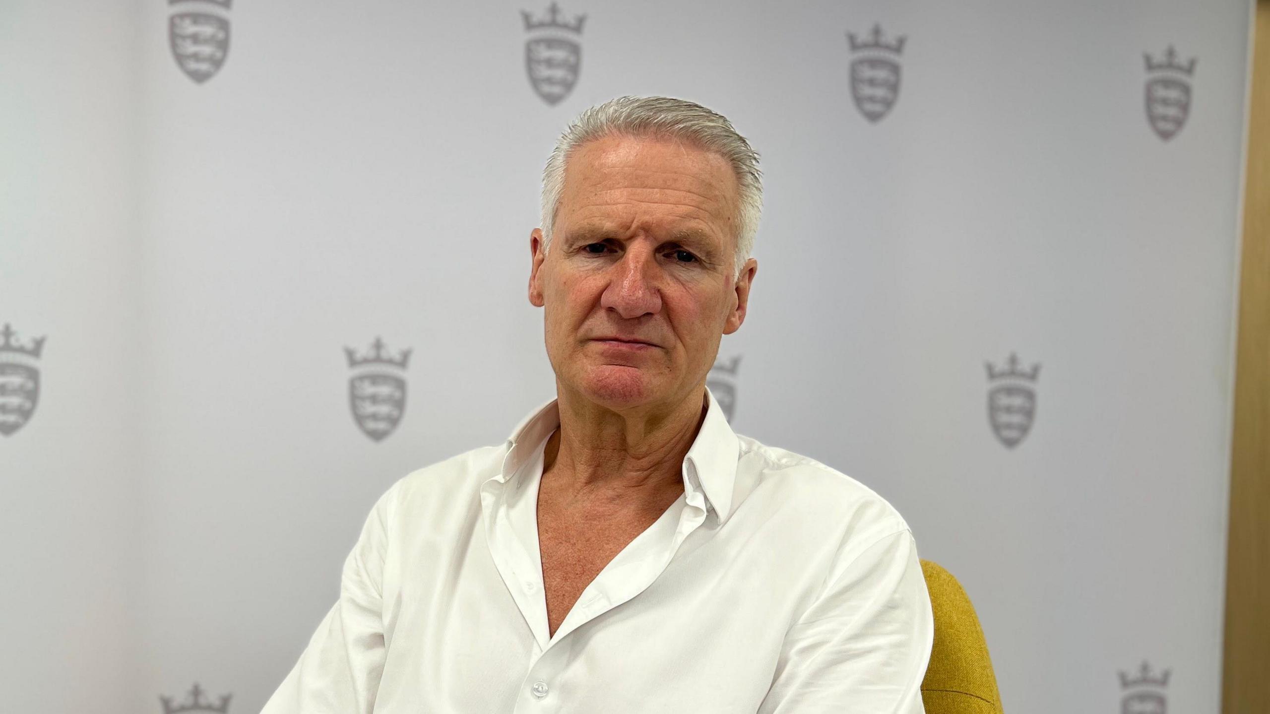 Tom Binet sits on a yellow chair in front of a backdrop featuring shield and crown logos. He is wearing a white shirt and blue jeans, looking directly at the camera.
