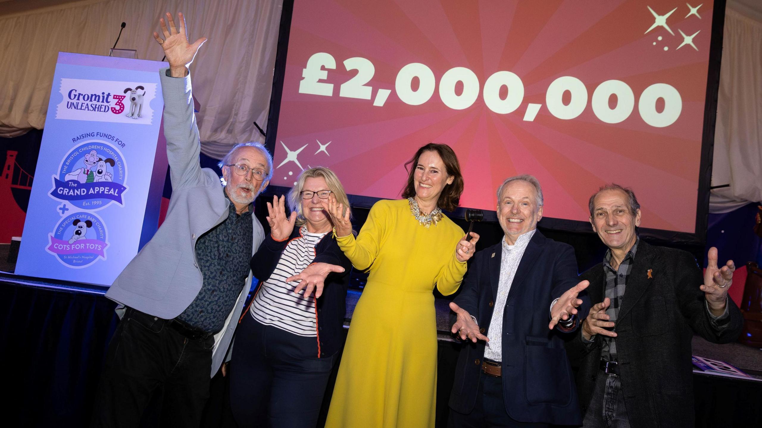 Five people wave their arms and smile as they face the camera. A woman in a yellow dress stands in the middle with a woman in a striped top and a man in a grey suit jacket stood to her. There are two men in suits on the other side of the woman in yellow. On one side of them is a banner publicising the Gromit Unleashed 3 auction with a light blue background. Above them is a large red screen with the figure £2,000,000 on it in thick red letters